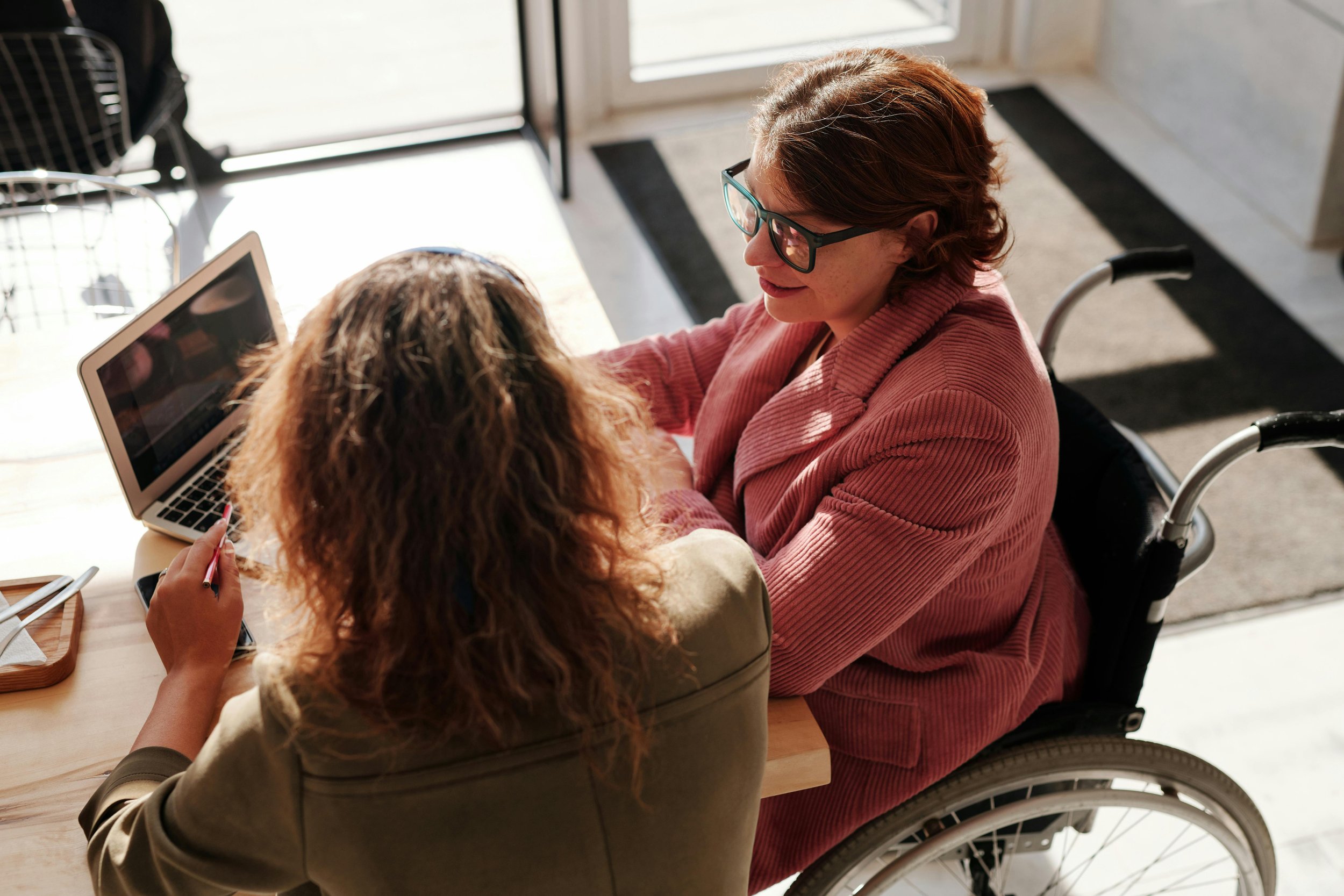 A woman in a wheelchair smiling and talking with another woman sitting at a desk with a laptop and a pen in her hand.