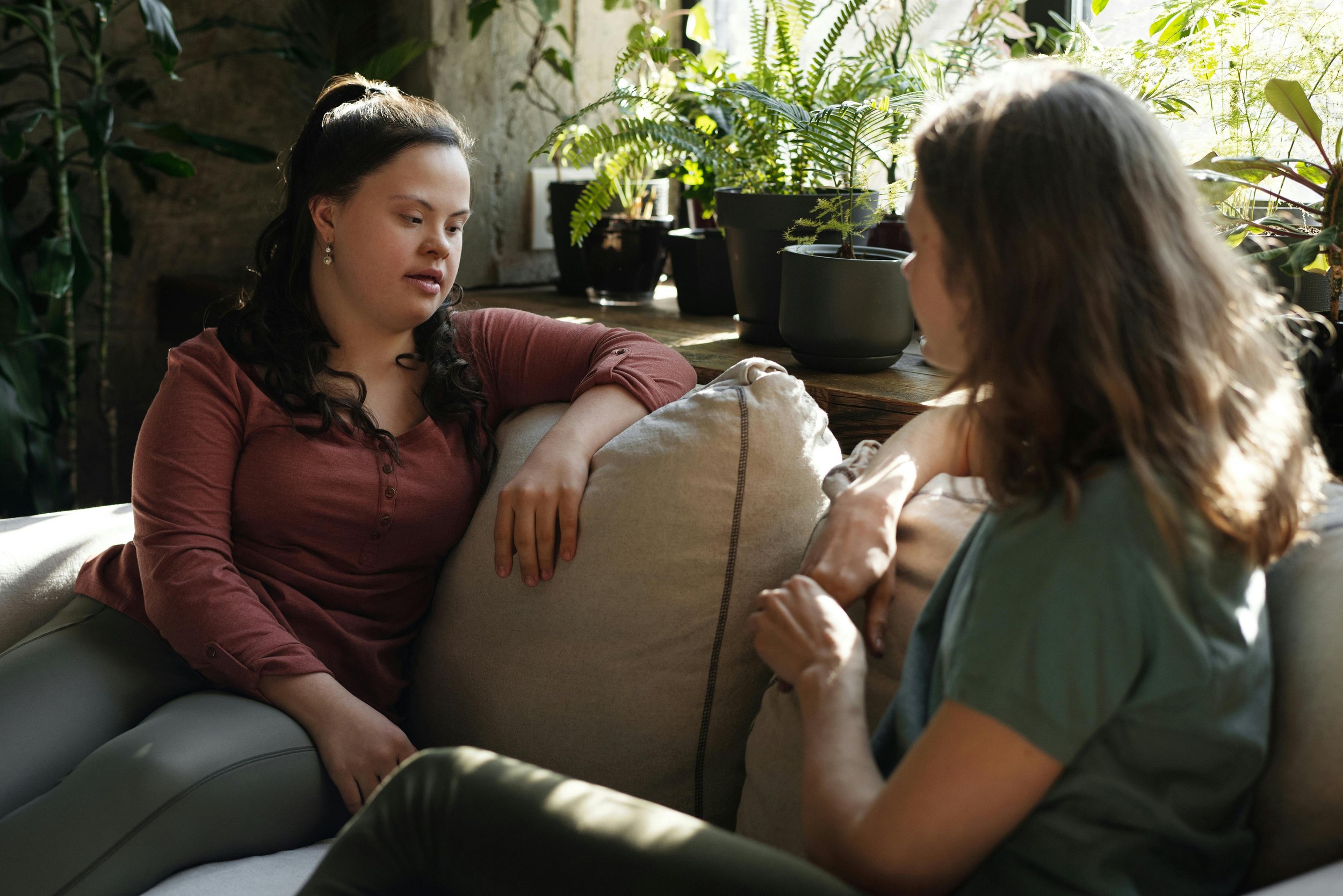 Two women are sitting on a sofa and engaging in a conversation. One woman has dark hair, is wearing a rust-colored top, and appears to be listening. The other woman has wavy brown hair, is wearing a green shirt, and is speaking. There are potted plants on a wooden surface behind them, and sunlight is coming through the window, creating a cozy atmosphere.