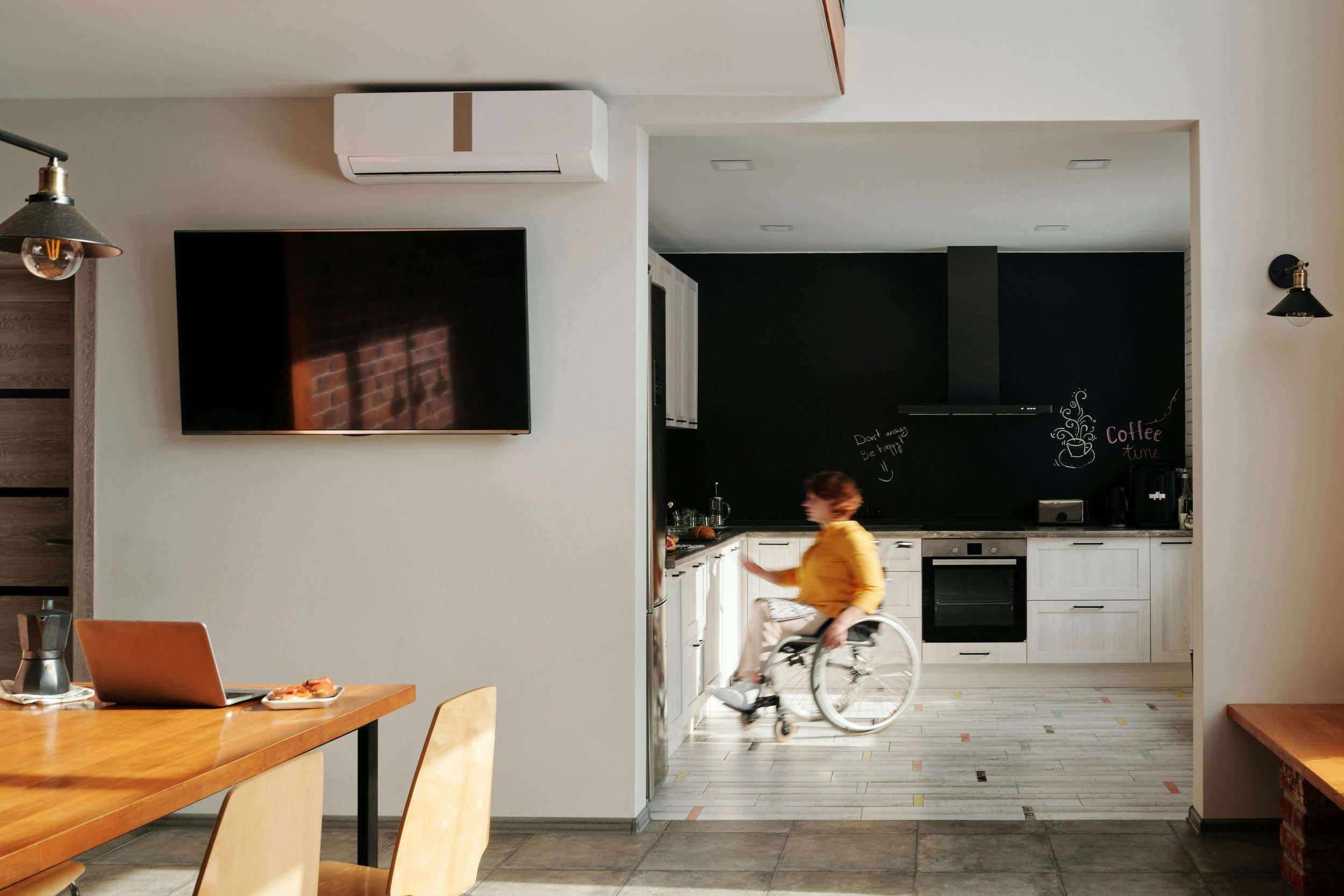 A woman in a yellow shirt in a wheelchair reaching into a kitchen with white cabinets and black backsplash, with a chalkboard wall that has writing and drawings, including a coffee mug drawing and the words "Coffee time". A TV is mounted on a white wall, and a dining table with chairs is partially visible in the foreground.