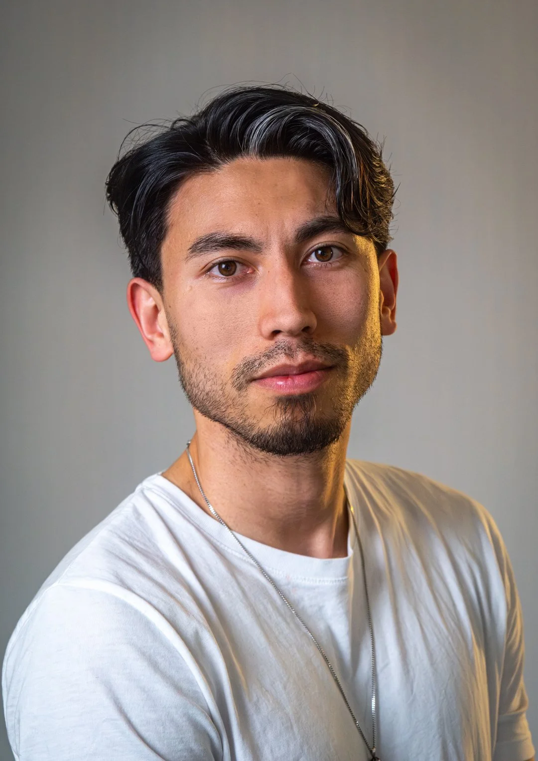 A young man with dark hair and a slight beard is looking directly at the camera with a neutral expression. He is wearing a white shirt and a thin necklace, set against a plain background.