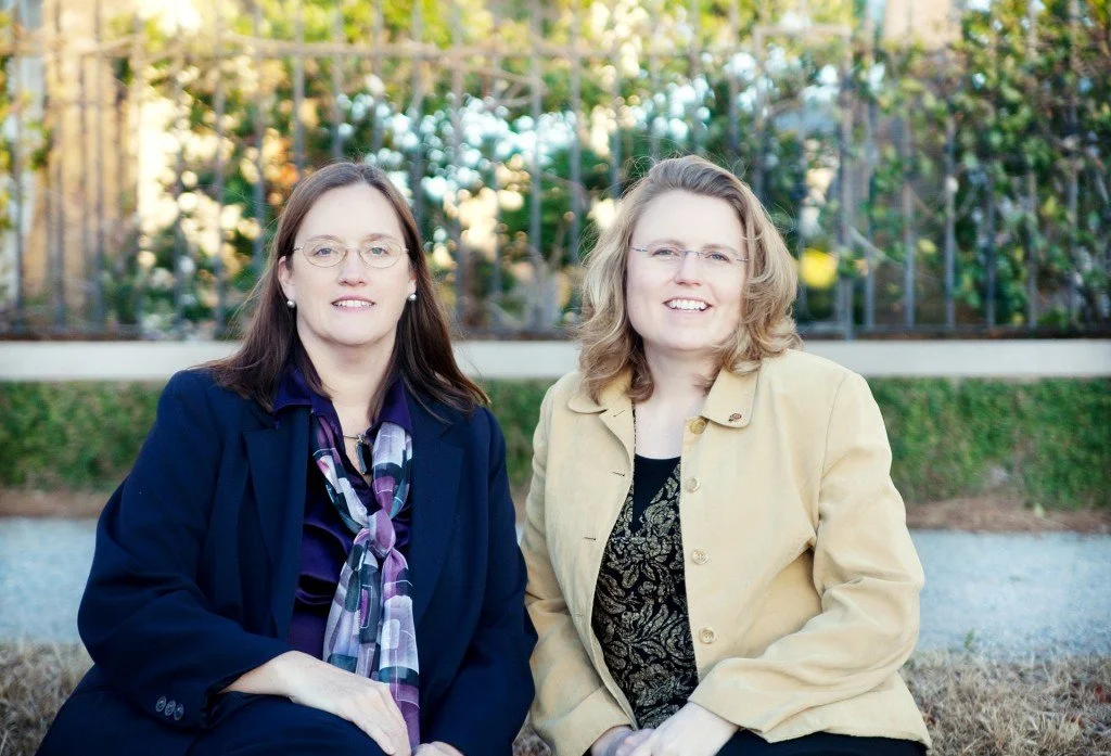 Two women sitting outdoors in front of a garden, smiling at the camera.