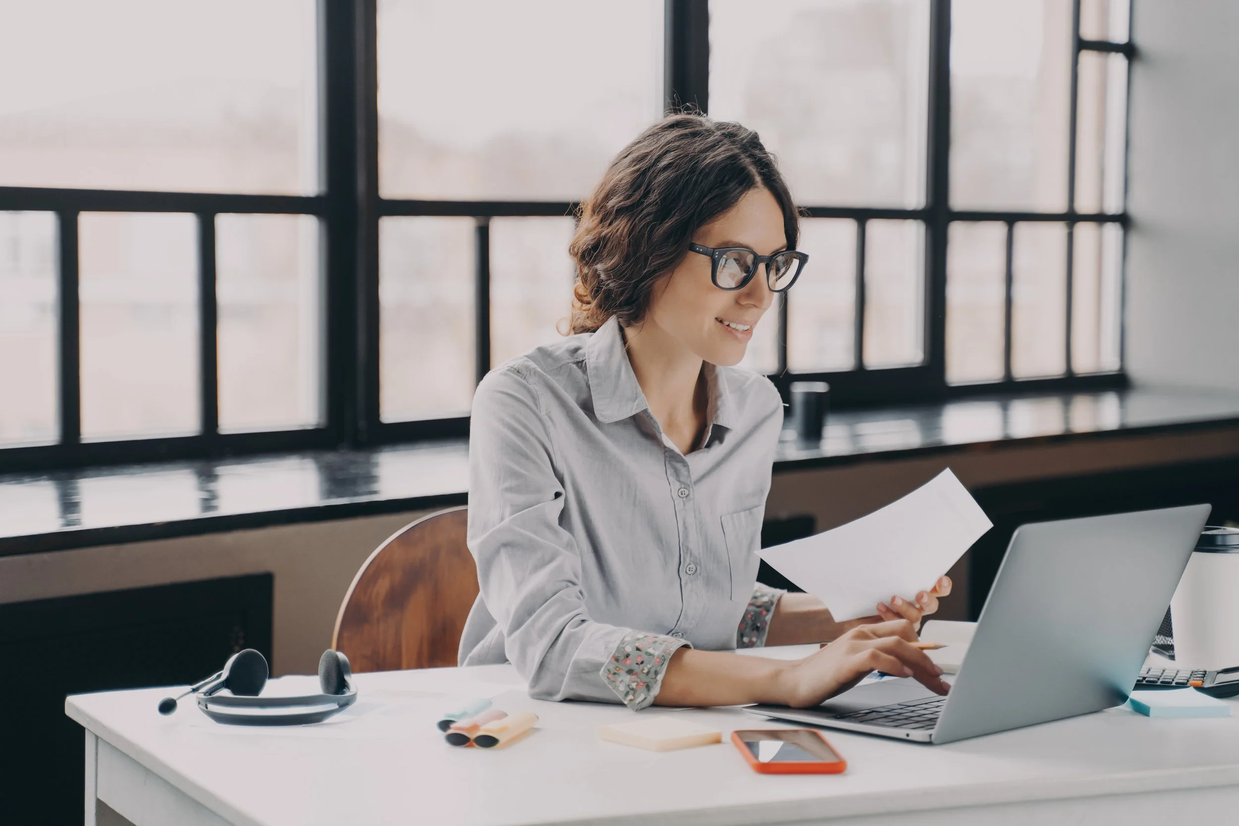 Woman with glasses working at a desk with a laptop, papers, and office supplies in a bright room with large windows.