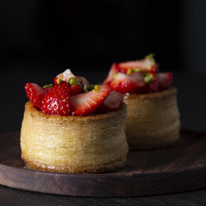 Zwei kleine Kuchen mit frischen Erdbeeren und Pistazien auf einem dunklen Holzbrett.