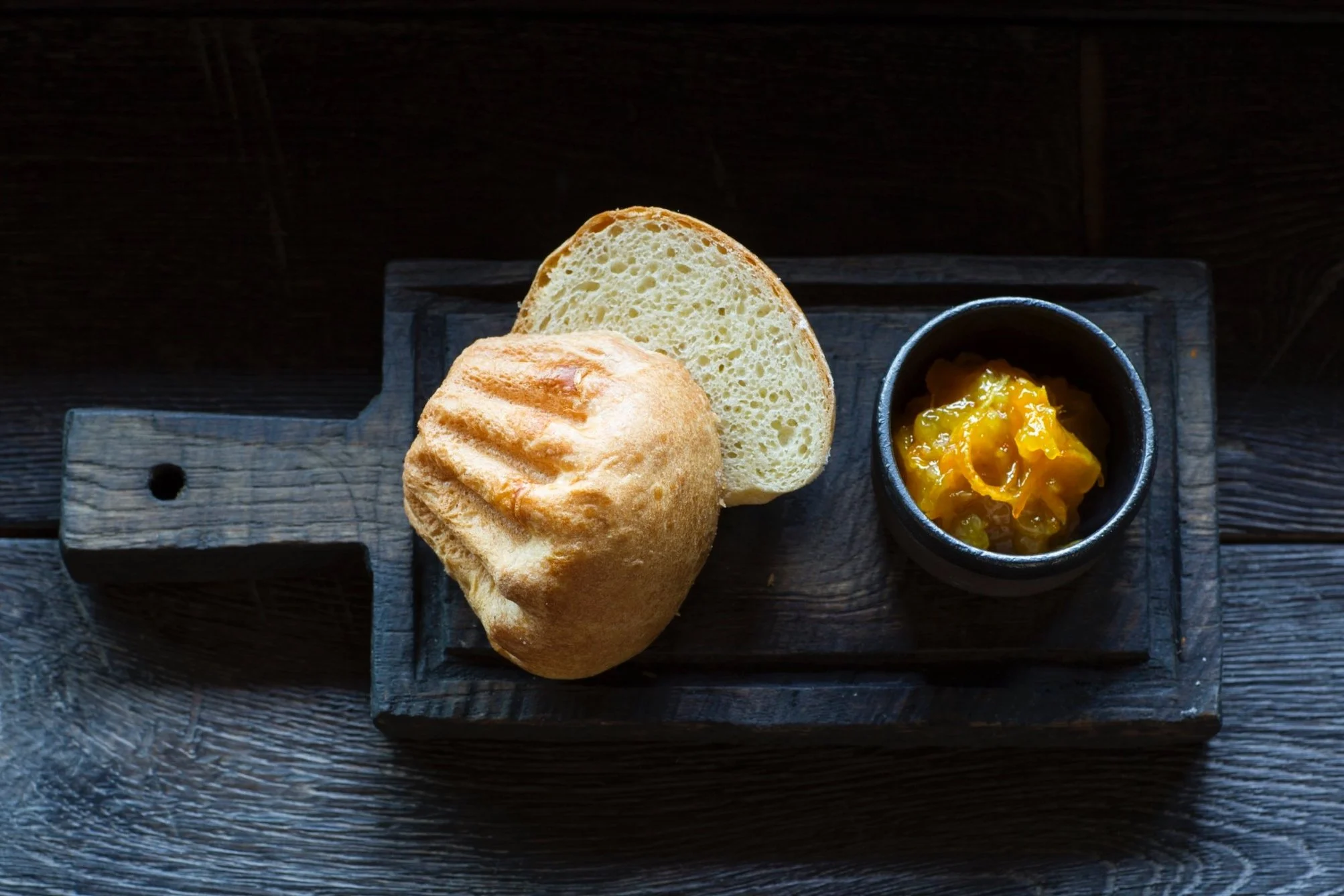 Brotscheibe, Brötchen und eine Schüssel mit Marmelade auf einem dunklen Holzbrett.