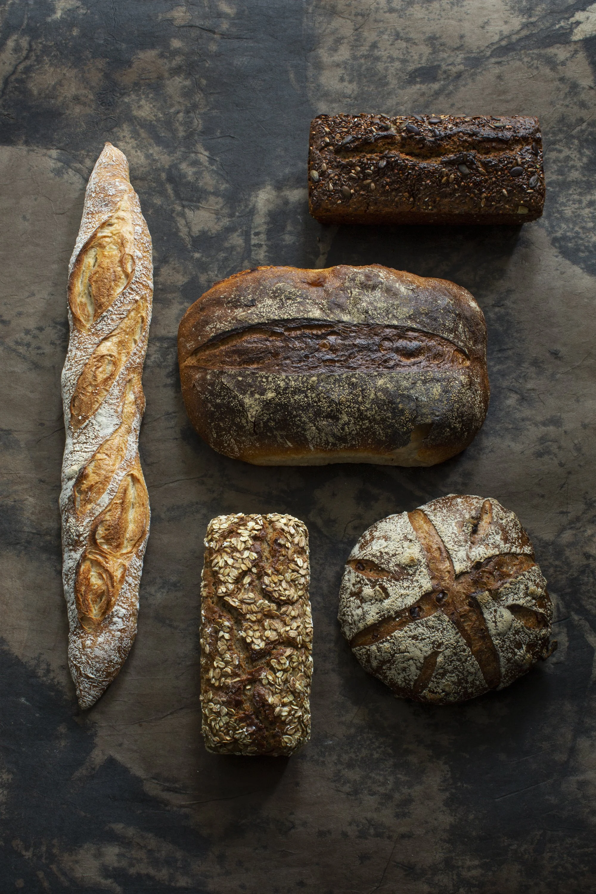 Verschiedene Brotsorten auf einem dunklen Steinboden, darunter ein Baguette, ein rustikales Brot mit Saaten, ein runder Brot mit Kreuzmustern, ein längliches dunkles Brot und ein ovales Brot mit Oberflächenstruktur.