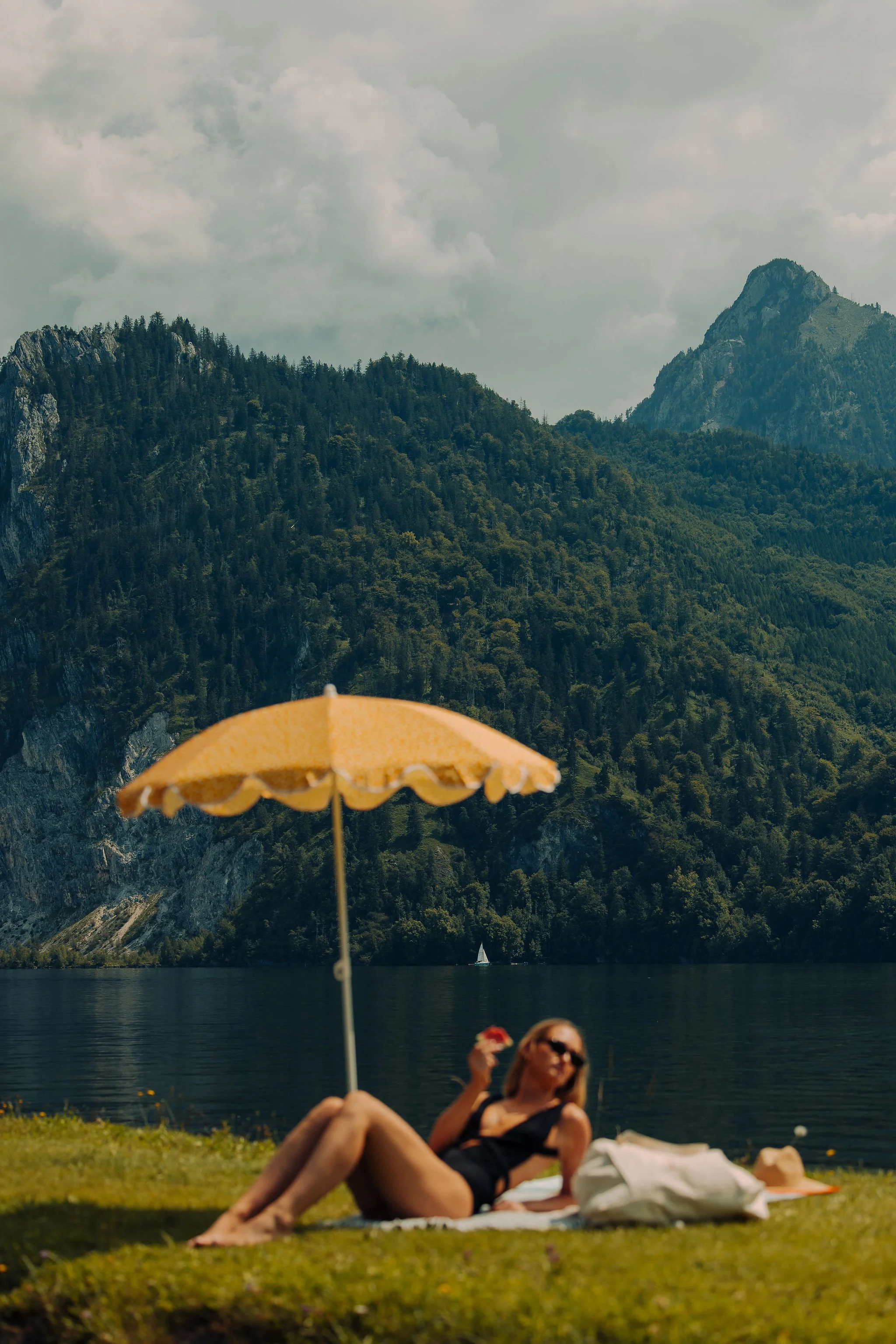 Eine Person liegt auf der Wiese unter einem Sonnenschirm in der Sonne, im Hintergrund sieht man Berge und den See. Inszeniert vom, Studio für Branding Doppelpaula.