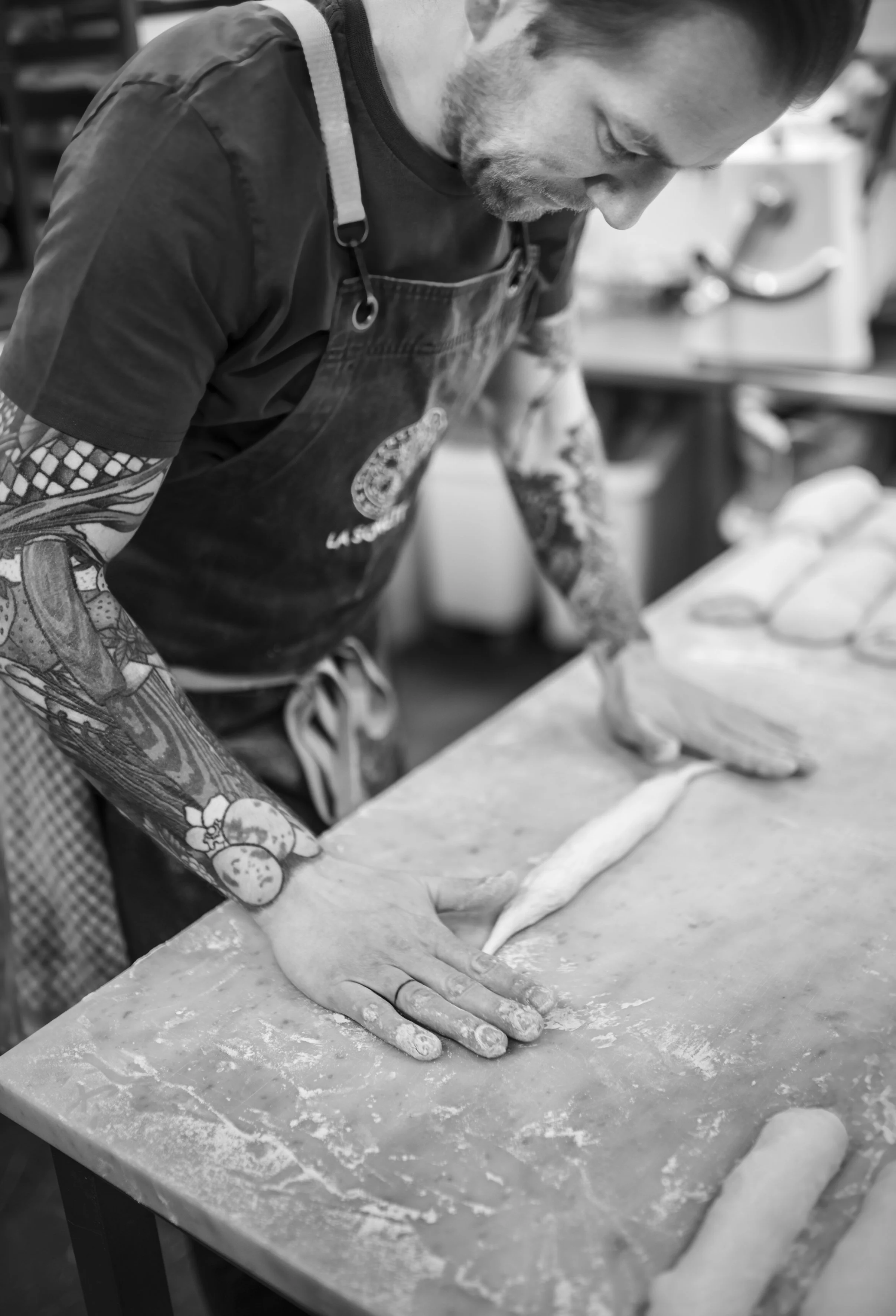 Ein tätowierter Mann bei der Zubereitung von Teig in einer Küche, er rollt Teig mit den Händen auf einer Arbeitsfläche aus.