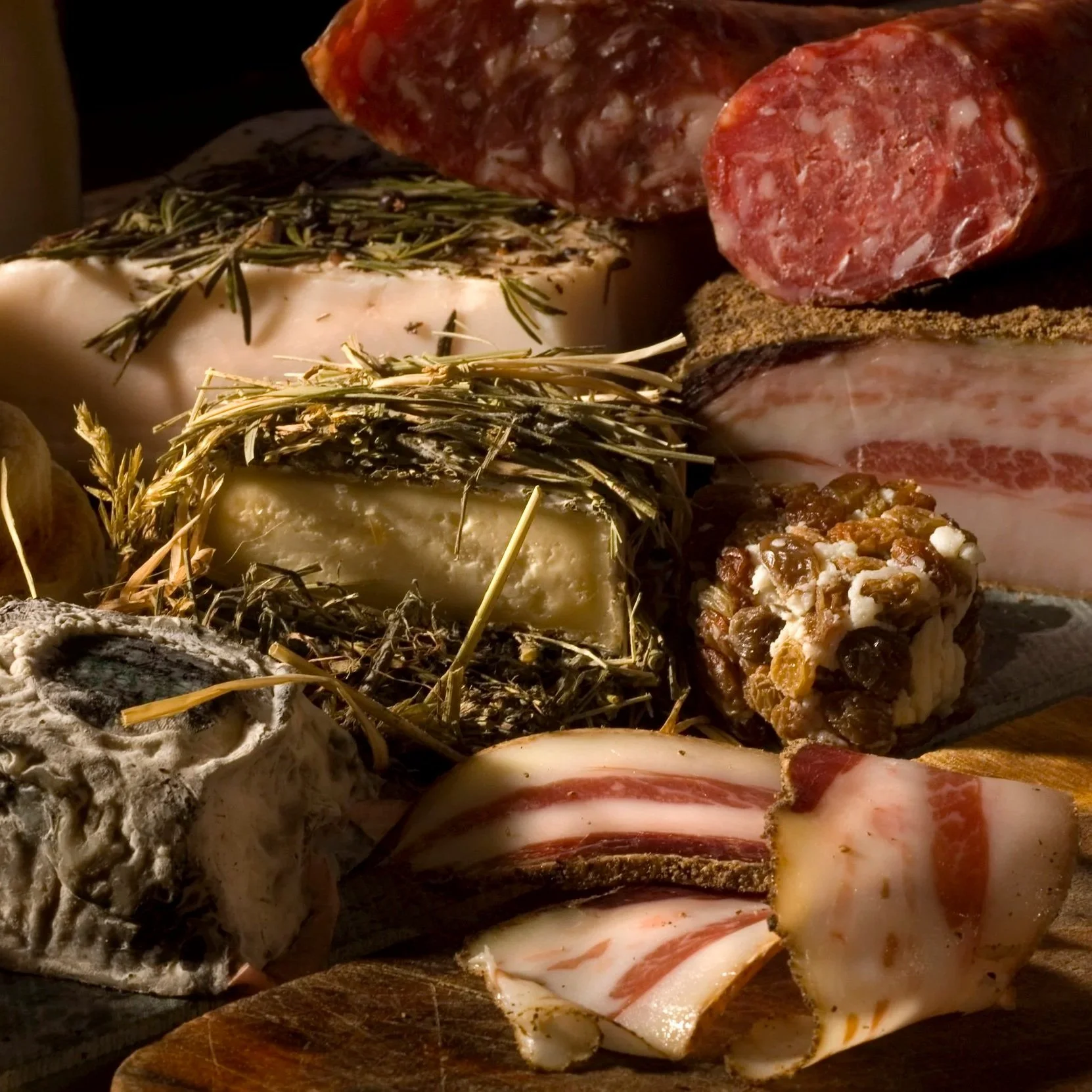Assorted cheeses and cured meats on a wooden board, including wedges of various cheese types, slices of bacon, and slices of salami.