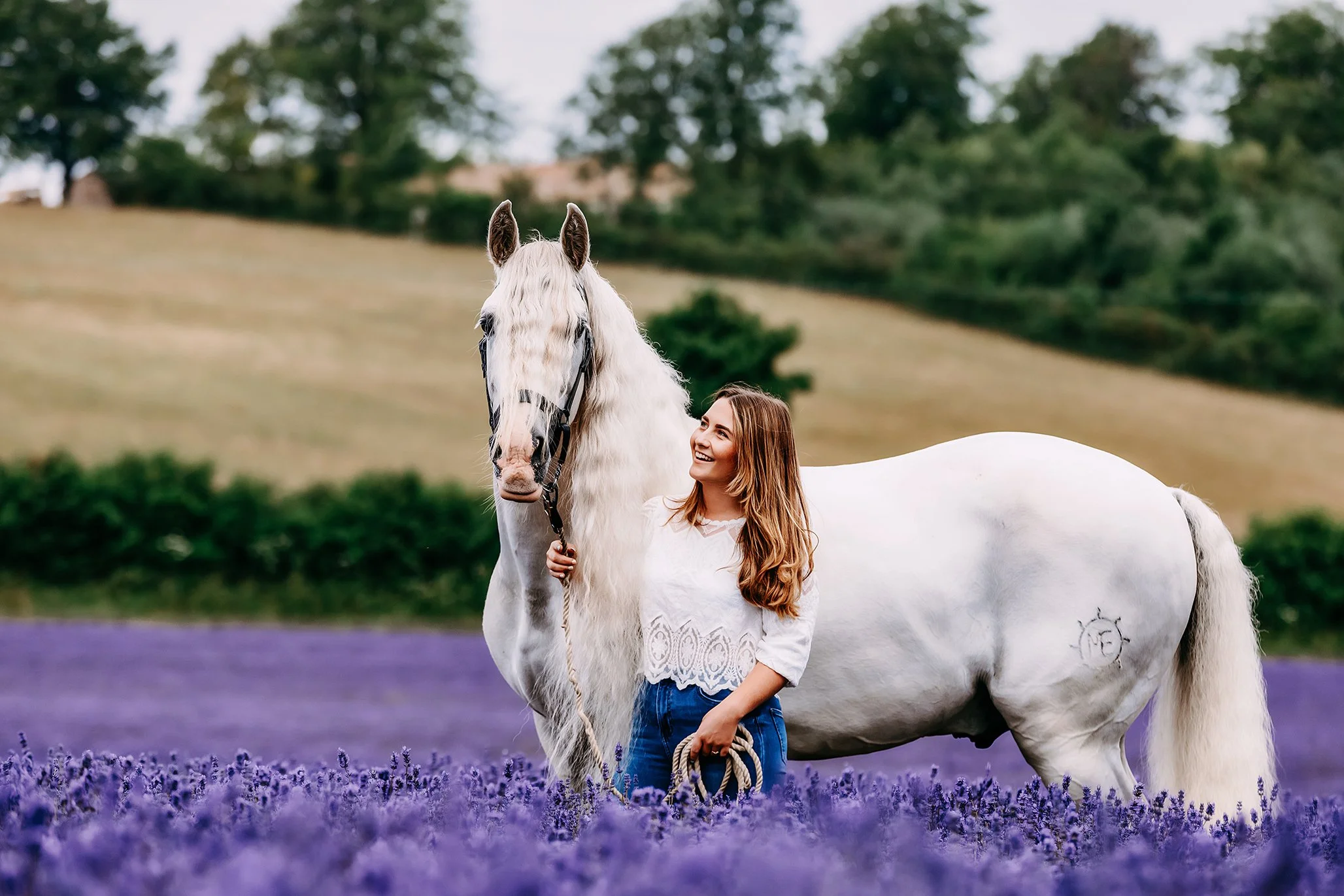 A woman standing next to a white horse in a lavender field, with a hillside and trees in the background.