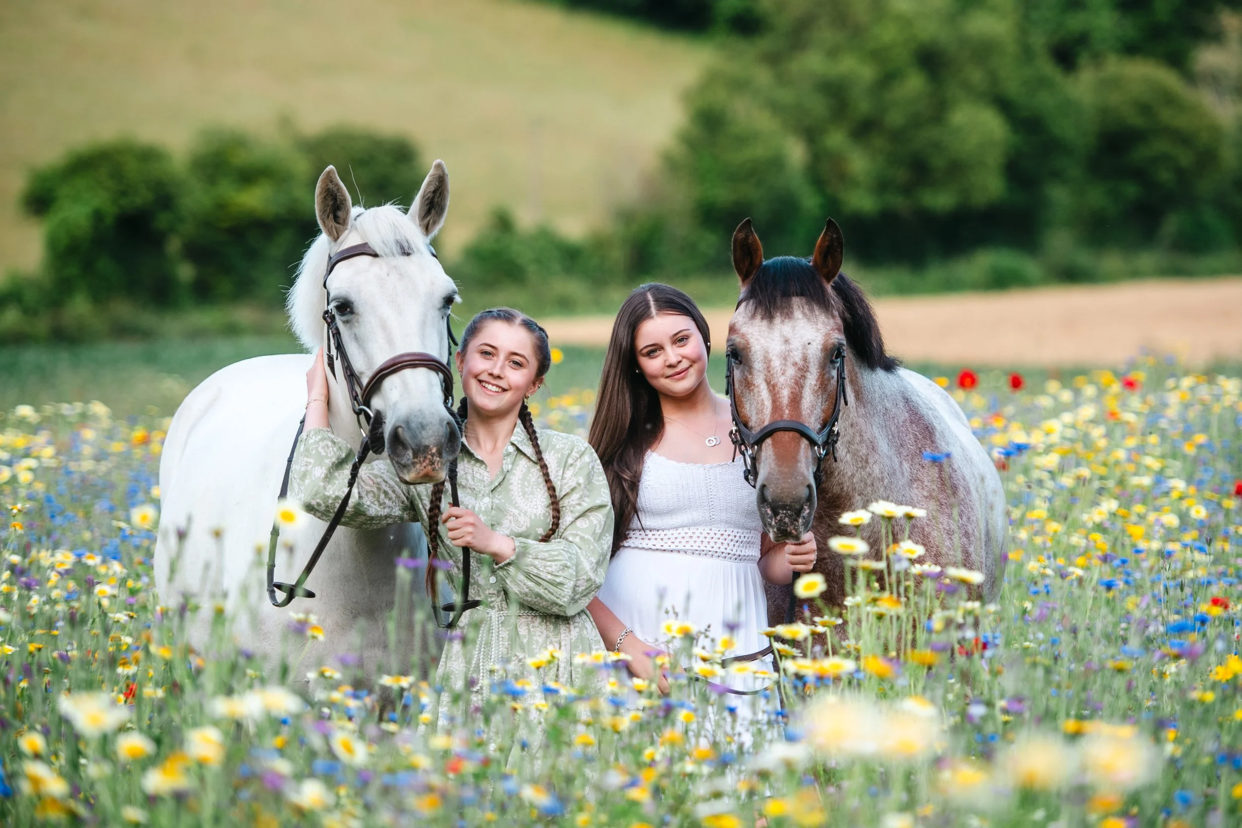Two young women standing in a field of wildflowers, each holding a horse on a lead. One woman is with a white horse, and the other with a gray horse. The background shows a lush green landscape.