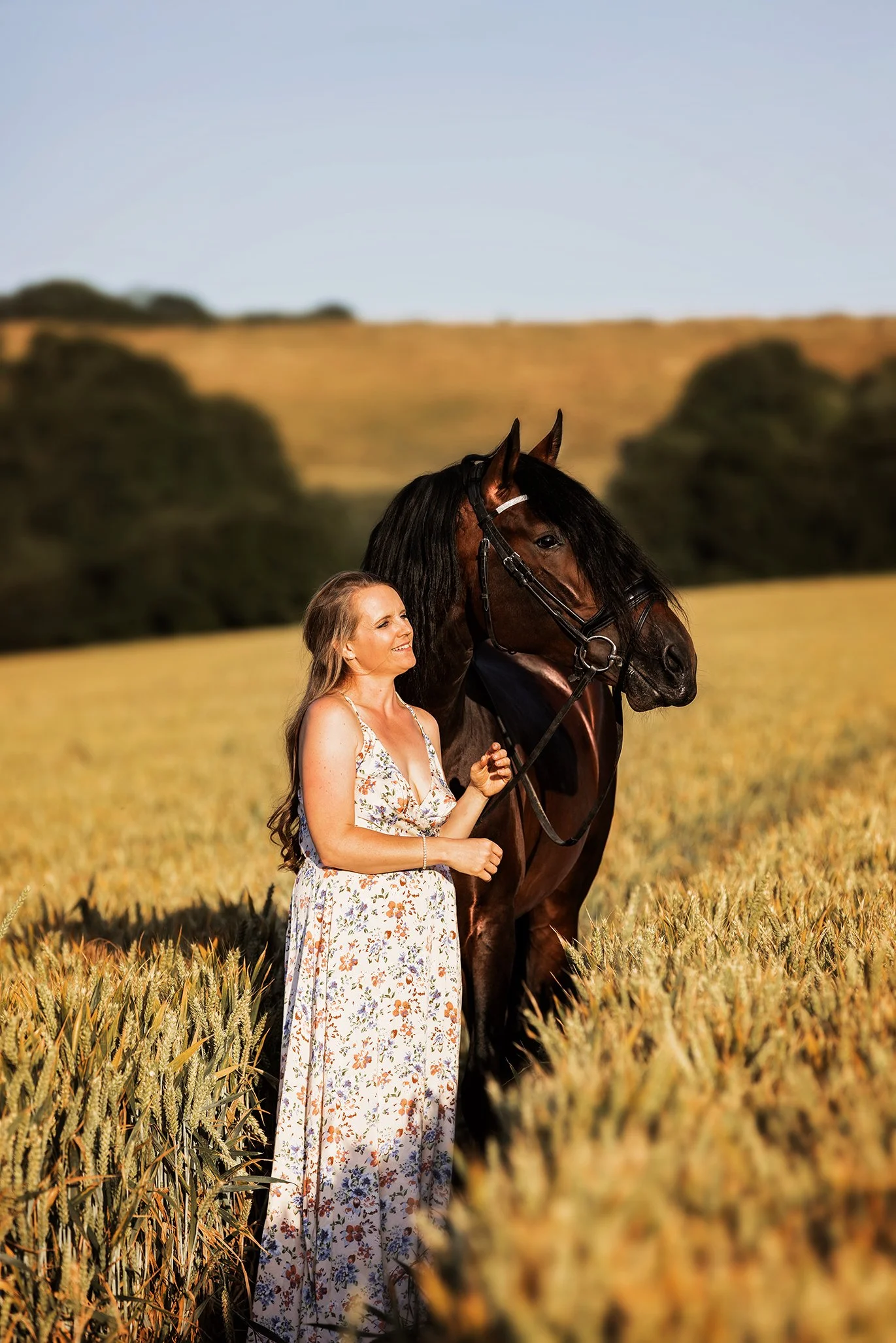 A woman in a long floral dress standing in a wheat field next to a large brown horse with a black mane during daytime.