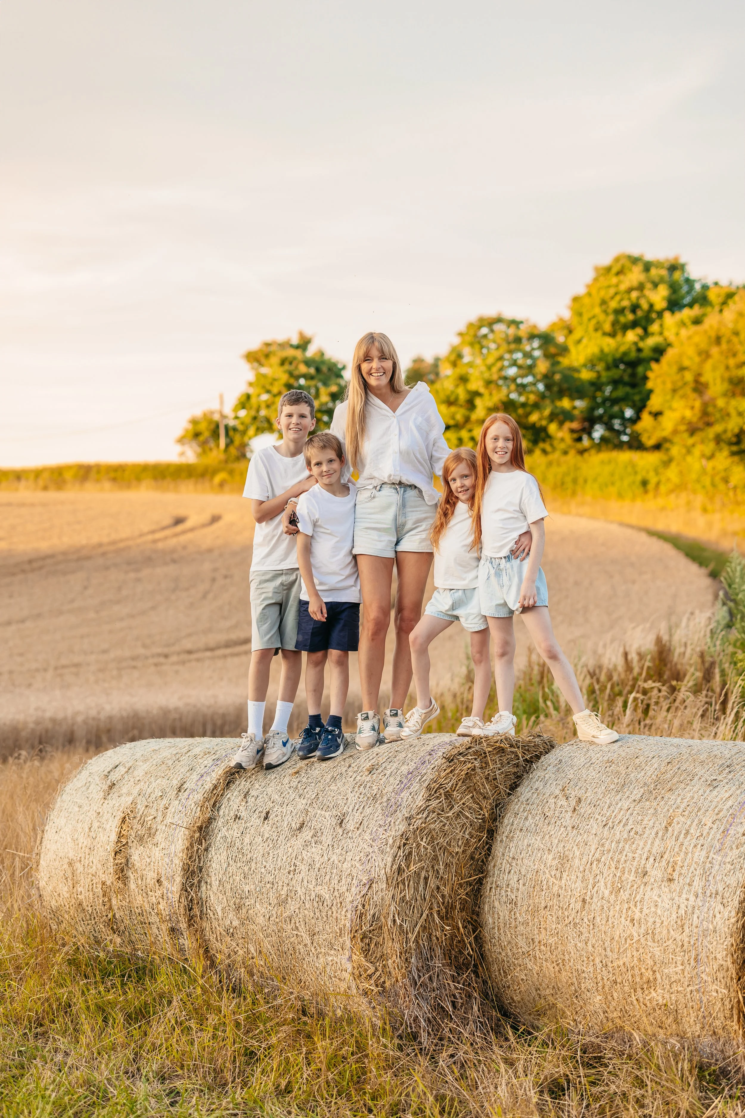 A smiling woman with four children standing on hay bales in a field during sunset, wearing casual white clothing.