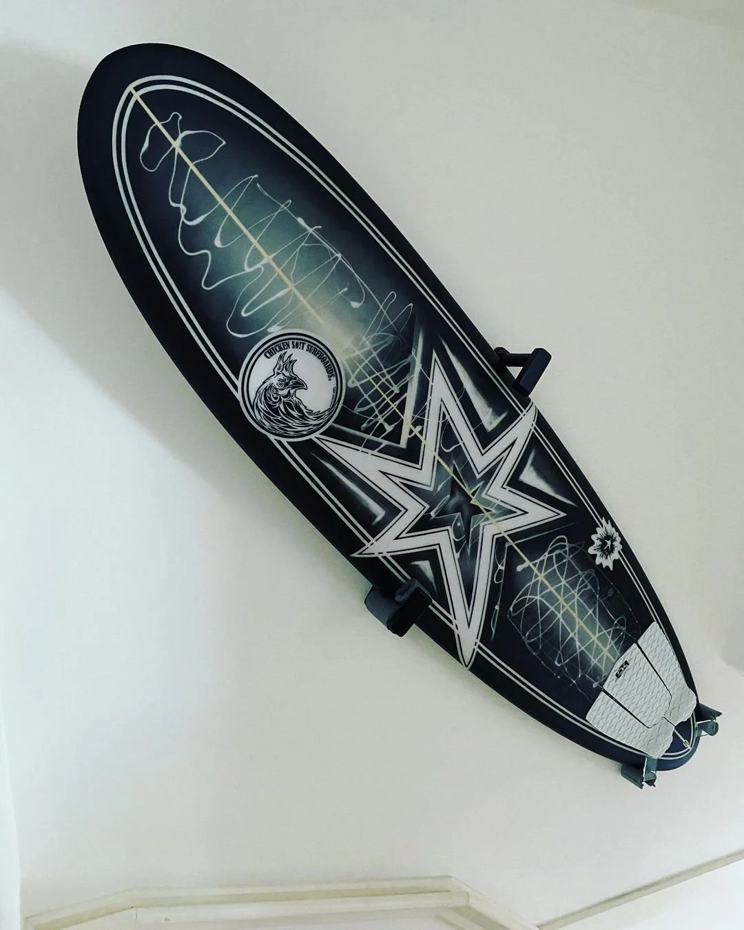 A black and white stand-up paddleboard with geometric and abstract designs, including a large star and a circle with a wolf illustration, mounted on a plain wall.