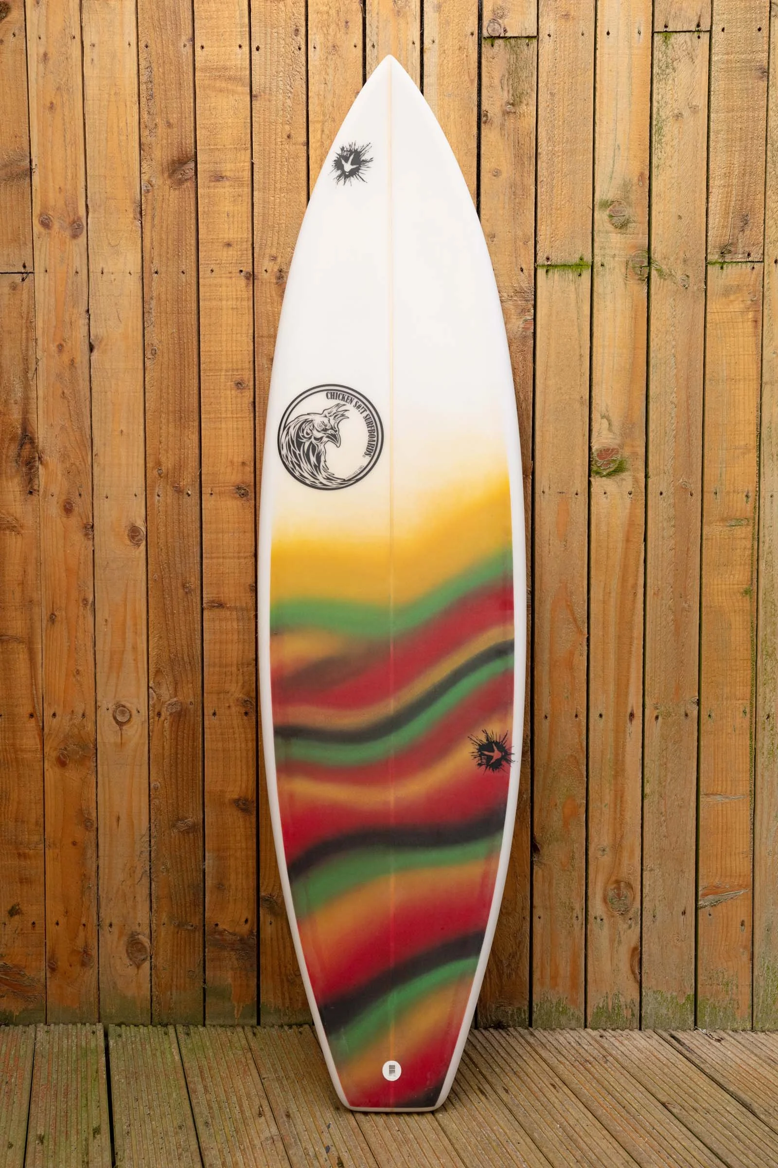 Colorful surfboard with a logo of a lion and the words 'Chicken Set-Free' leaned against a wooden fence.