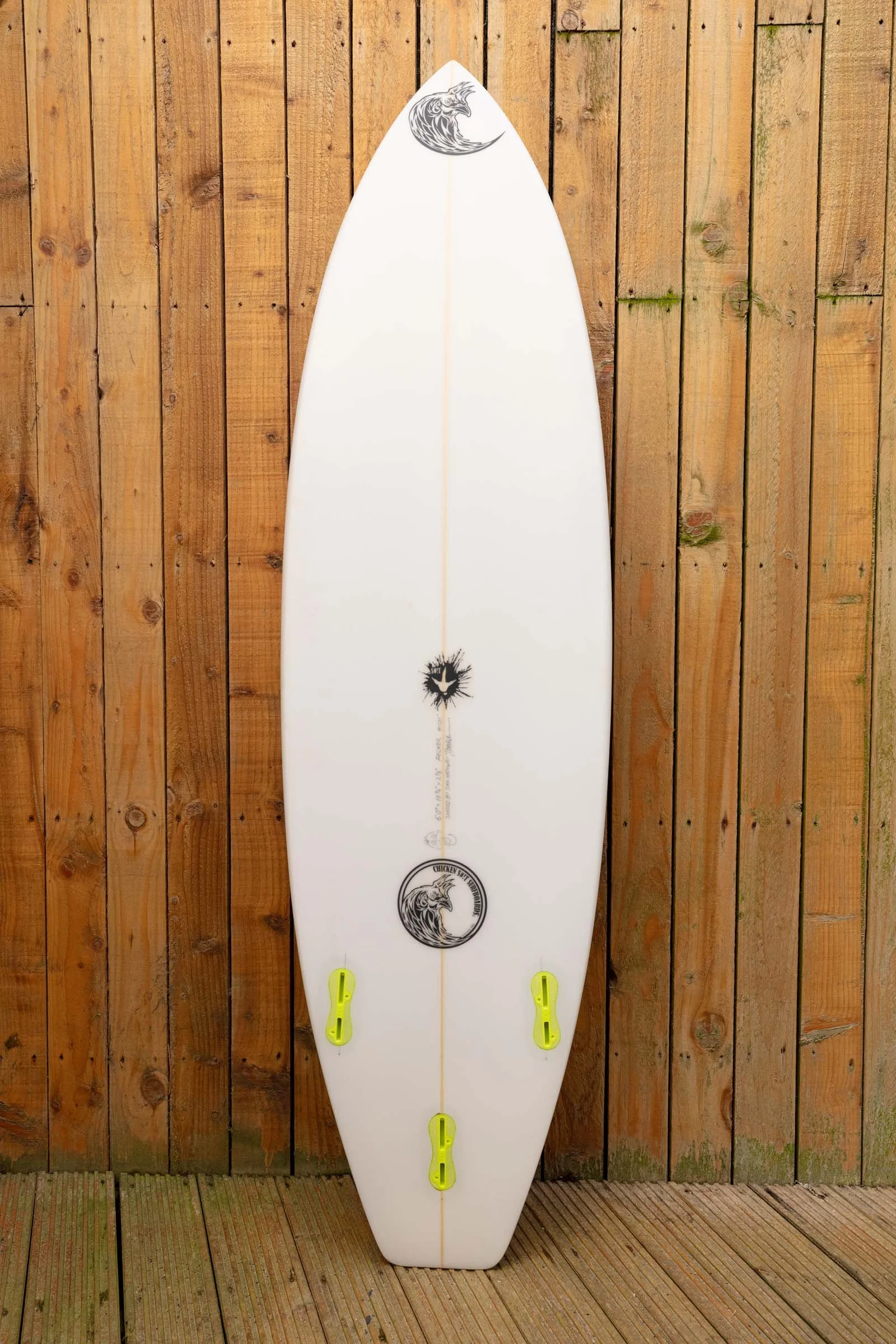 A white surfboard with a black and white wave logo at the top, black and white logo towards the bottom, and neon yellow fins, leaning against a wooden fence.