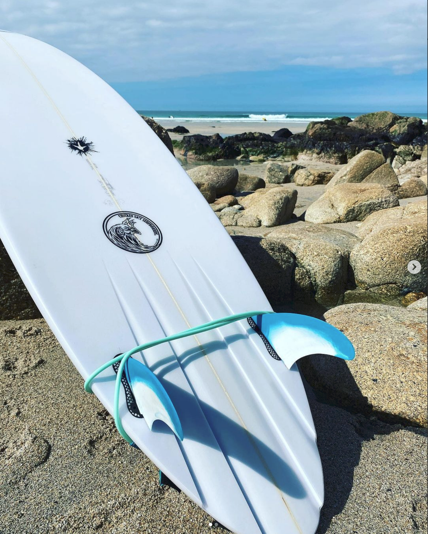 A white surfboard with blue fins resting on sandy beach near rocks, with ocean waves and a cloudy sky in the background.