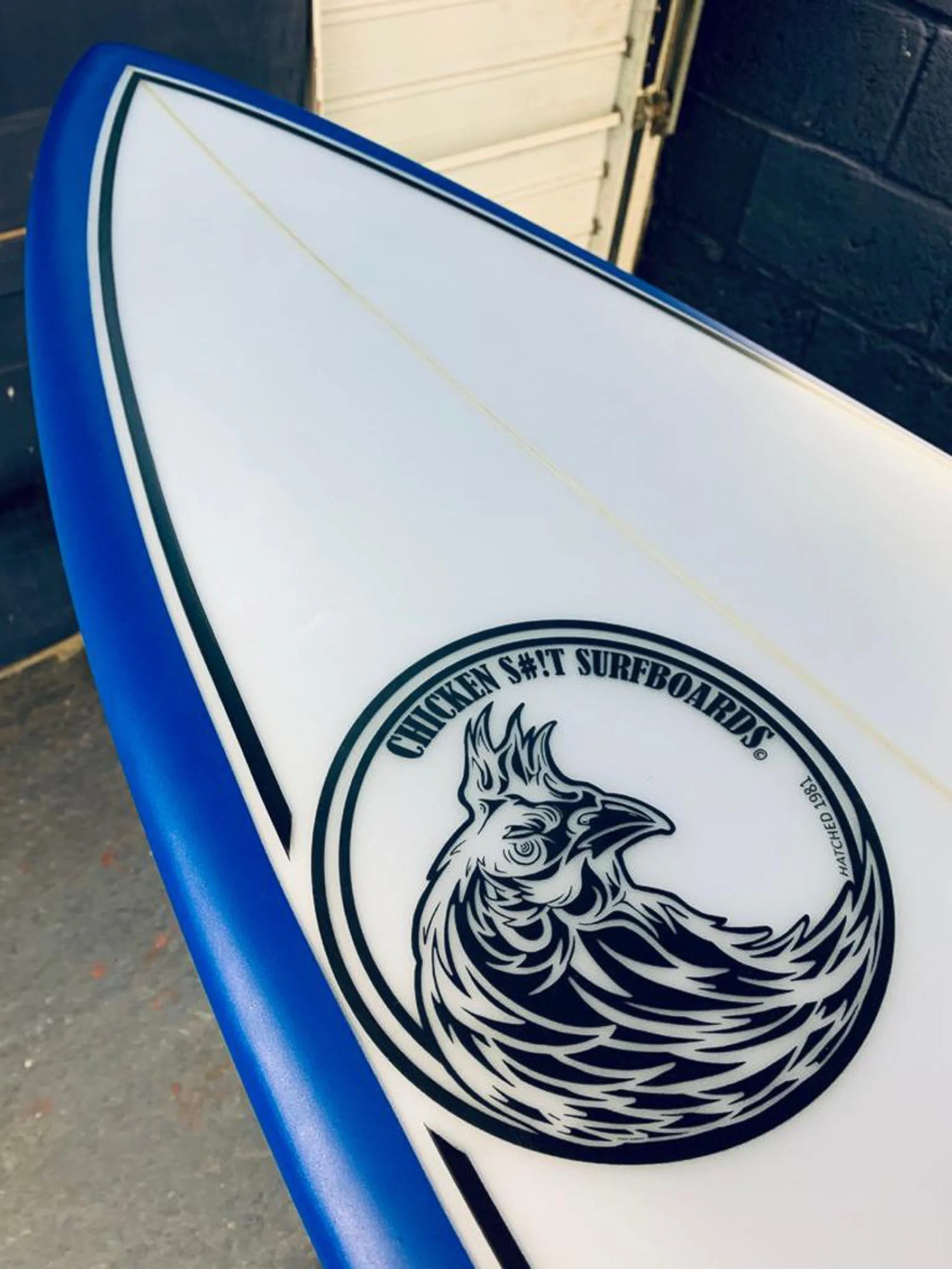 Close-up of a surfboard with a logo that reads 'Chicken Sh!t Surfboards' and features a stylized rooster design.