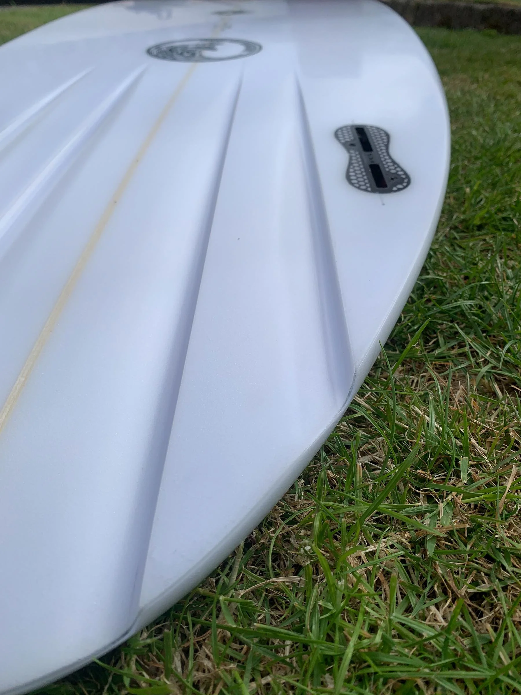 Close-up of a white surfboard on grass with fins and a leash attached.