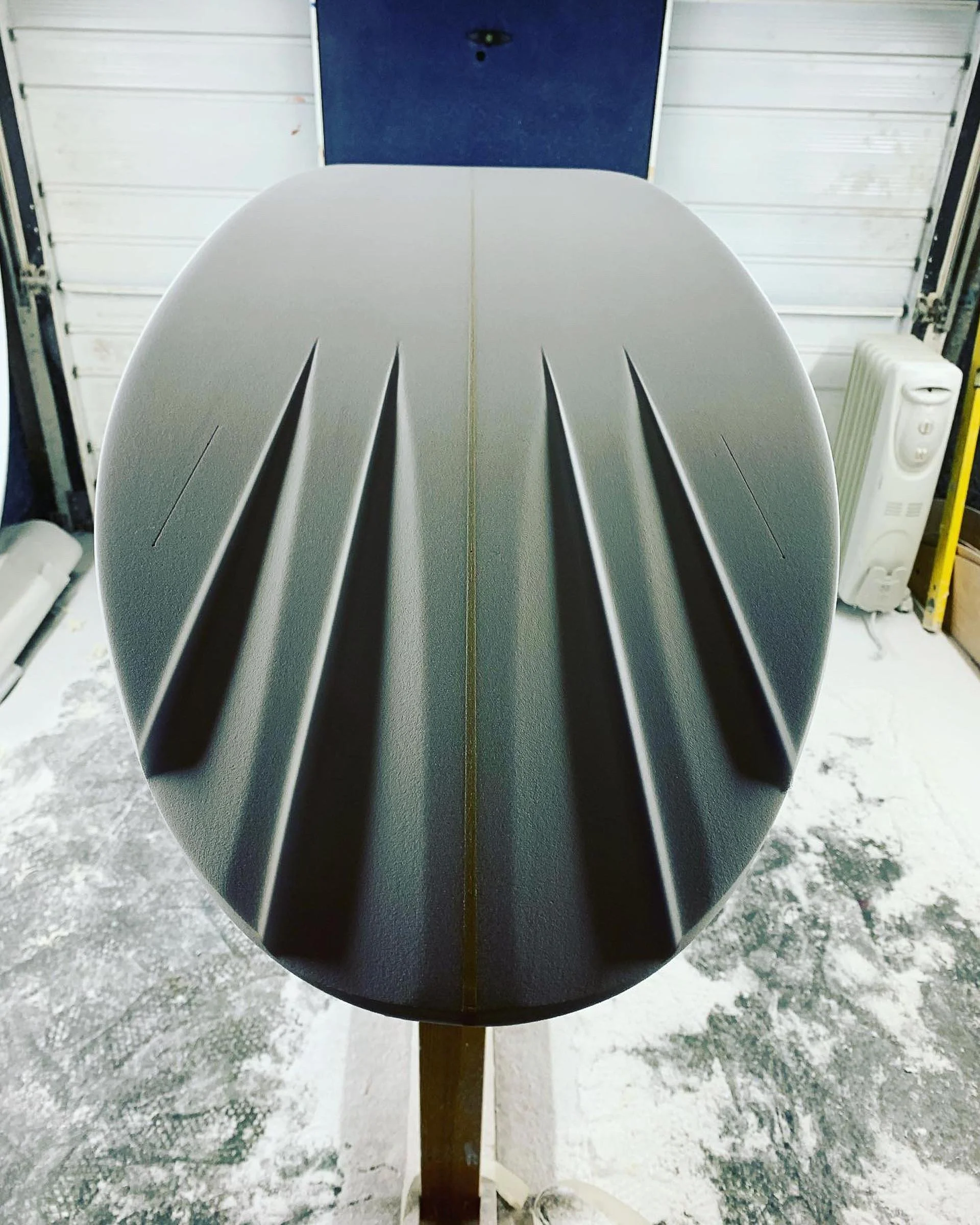 The image shows the top view of a surfboard with three long, black fins attached to the bottom, placed in a garage or workshop setting with a blue door, a heater, and some tools visible in the background.