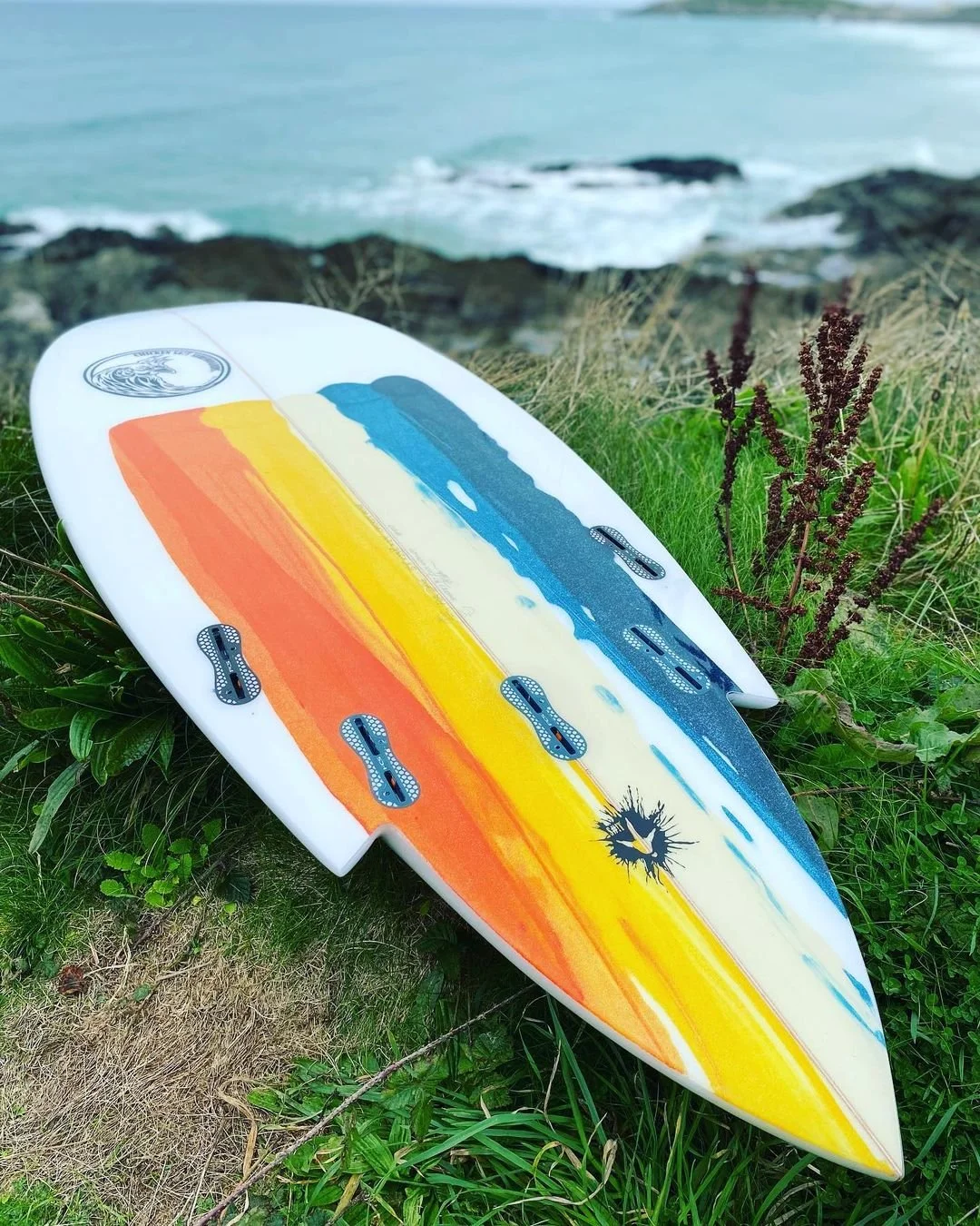 A colorful surfboard with a rainbow design resting on grassy ground near the ocean with rocks and waves.