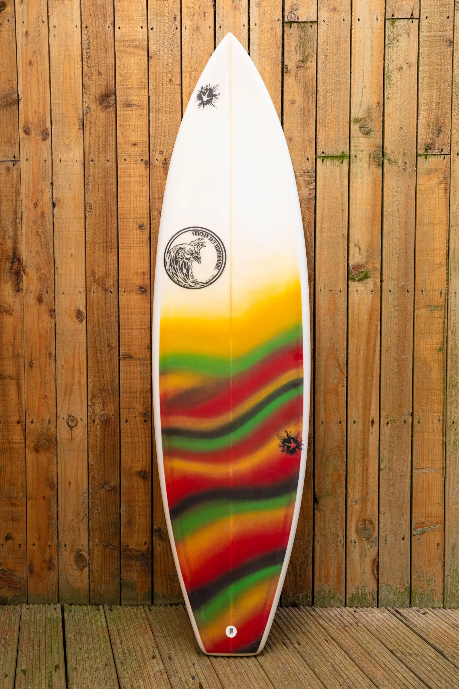 A colorful surfboard with a rainbow pattern leaning against a wooden fence.