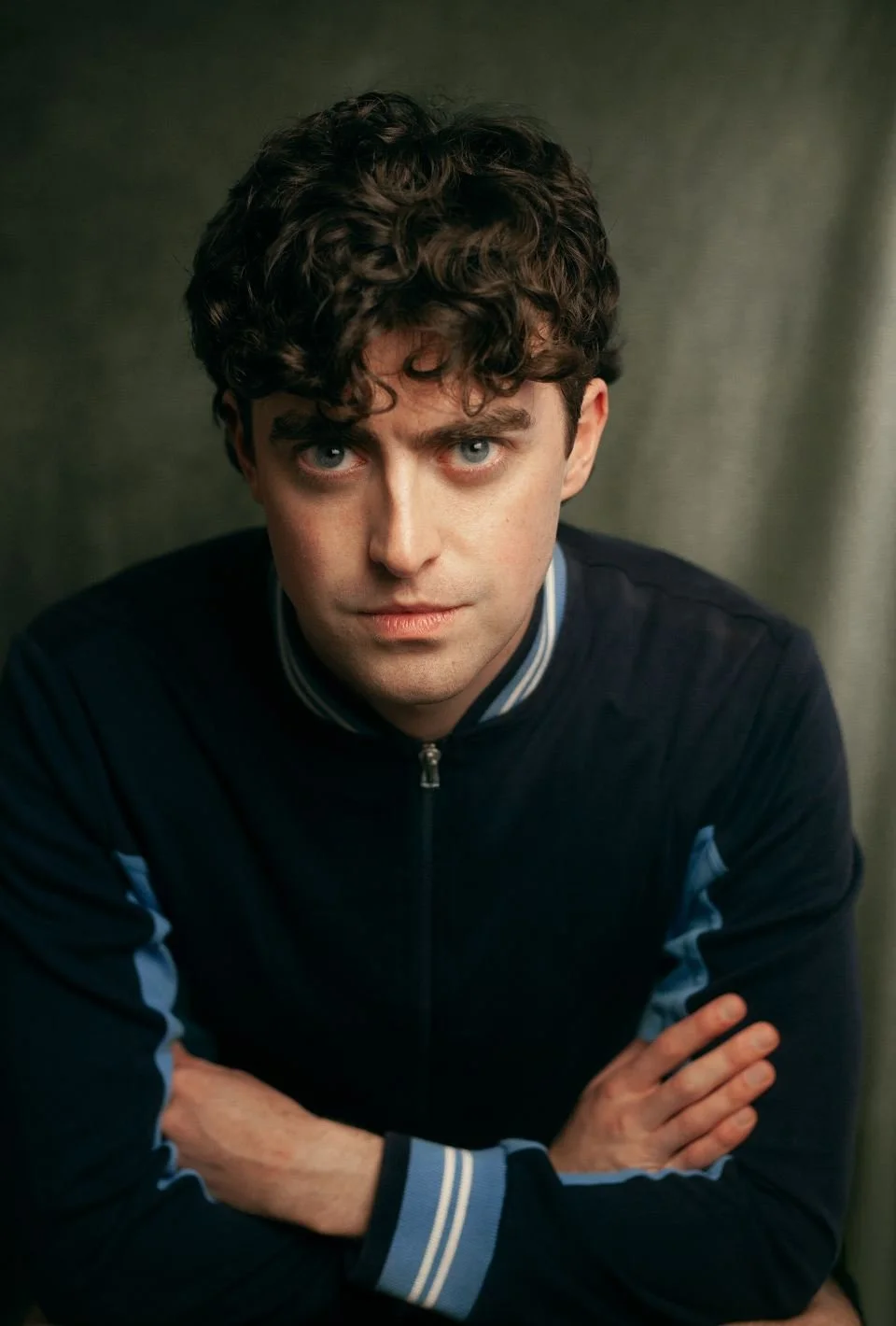 A young man with curly brown hair and blue eyes, wearing a dark blue zip-up jacket with light blue and white striped cuffs, looking intently at the camera with arms crossed, against a neutral background.