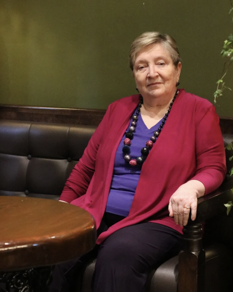 An older woman sitting on a black leather booth in a restaurant, wearing a red jacket, purple top, and a multi-colored beaded necklace.