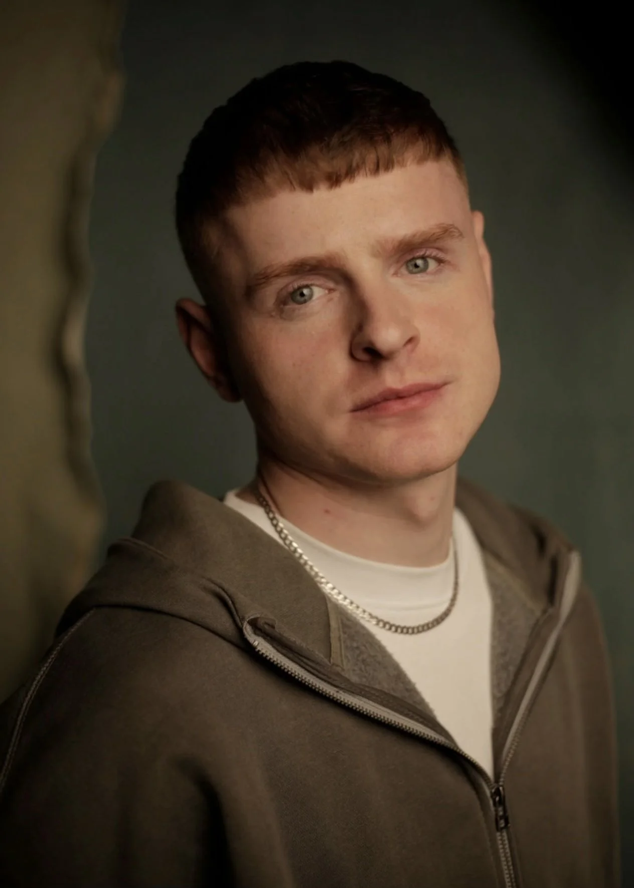 Portrait of a young man with short red hair, blue eyes, and earrings, wearing a black sweater with a white collar, against a blurred gray background.
