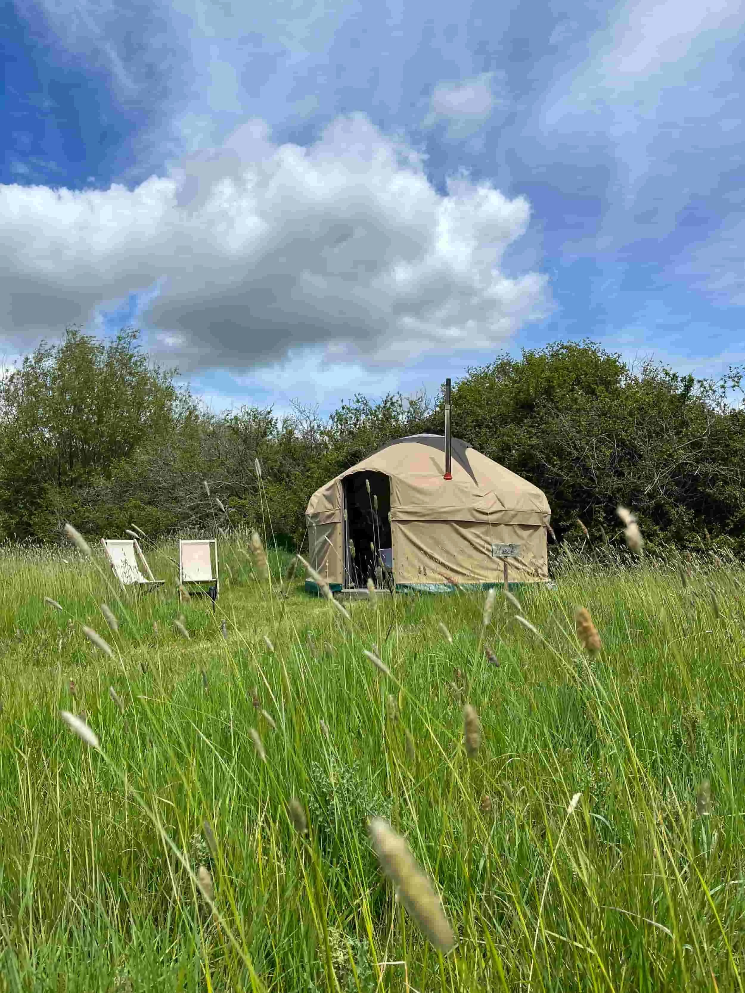 Handbuilt yurt in long meadow grass with deckchairs, dramatic cloud sky behind, Botswicky Wiltshire