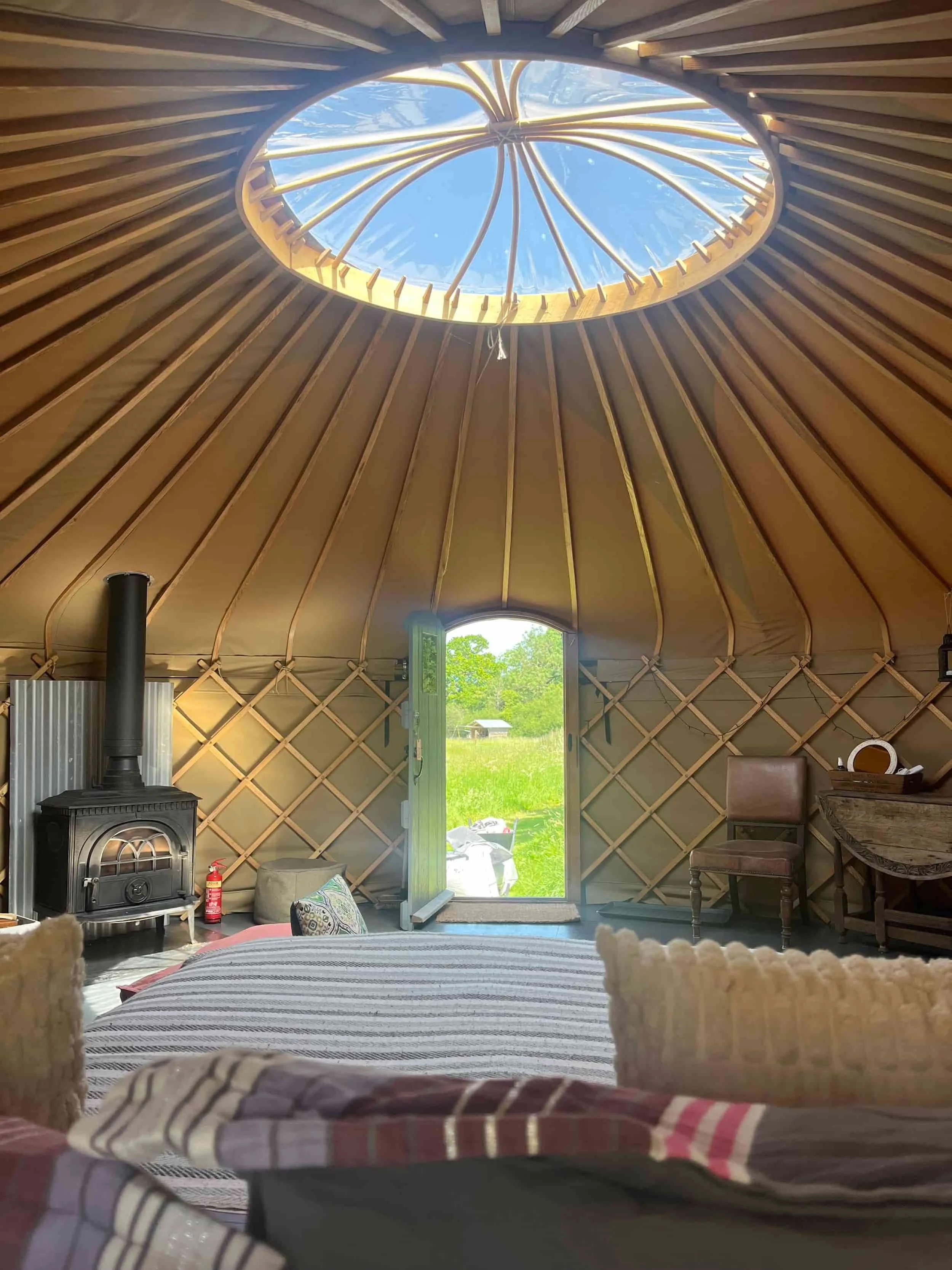 nside a handbuilt yurt — wood-burning stove, crown wheel above, open door framing a view across the meadow, Botswicky