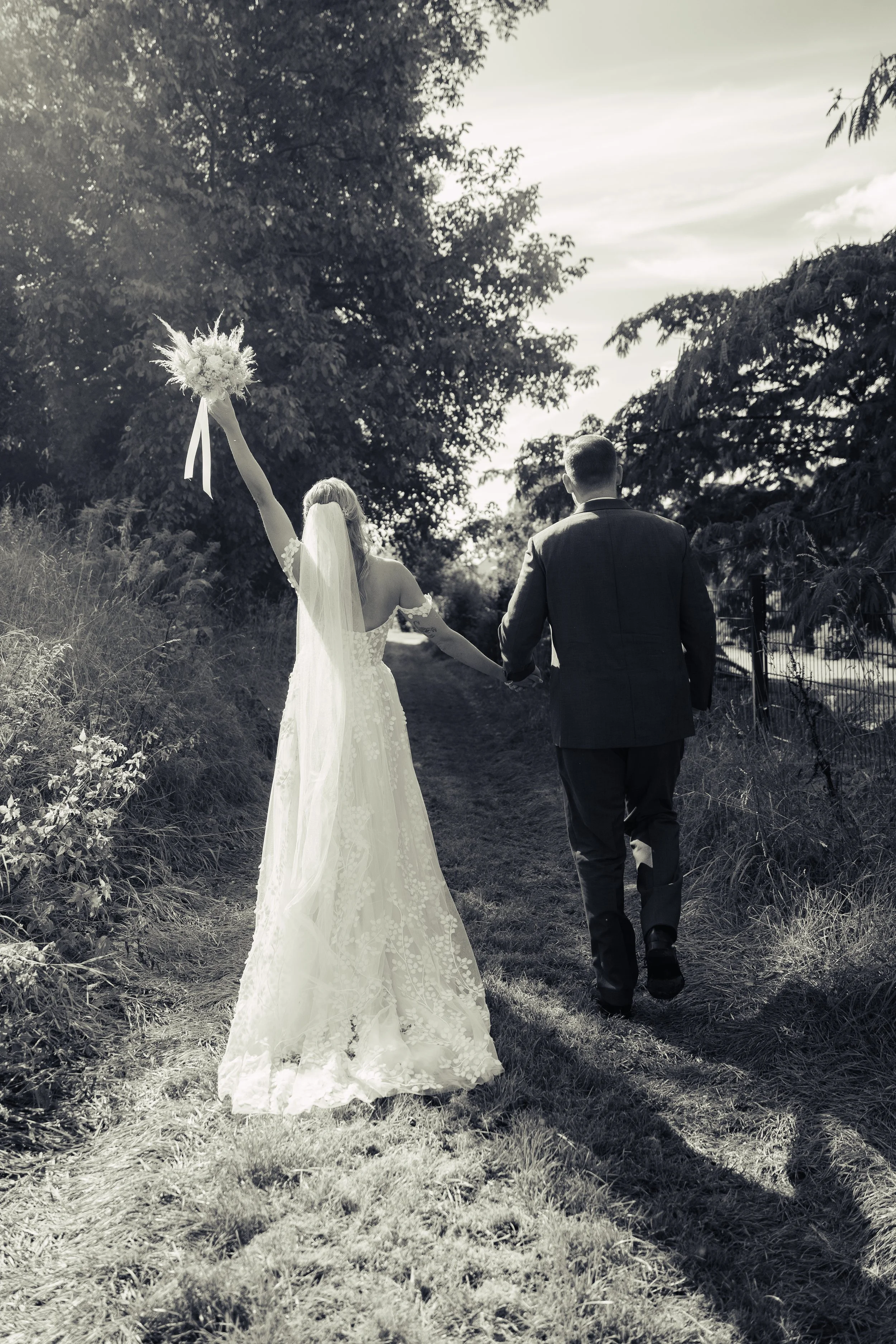 Braut und Bräutigam gehen Hand in Hand auf einem Feldpfad, die Braut hält ein Blumenbouquet hoch, es ist schwarz-weiß Fotografie, sonniger Tag, umgeben von Bäumen.