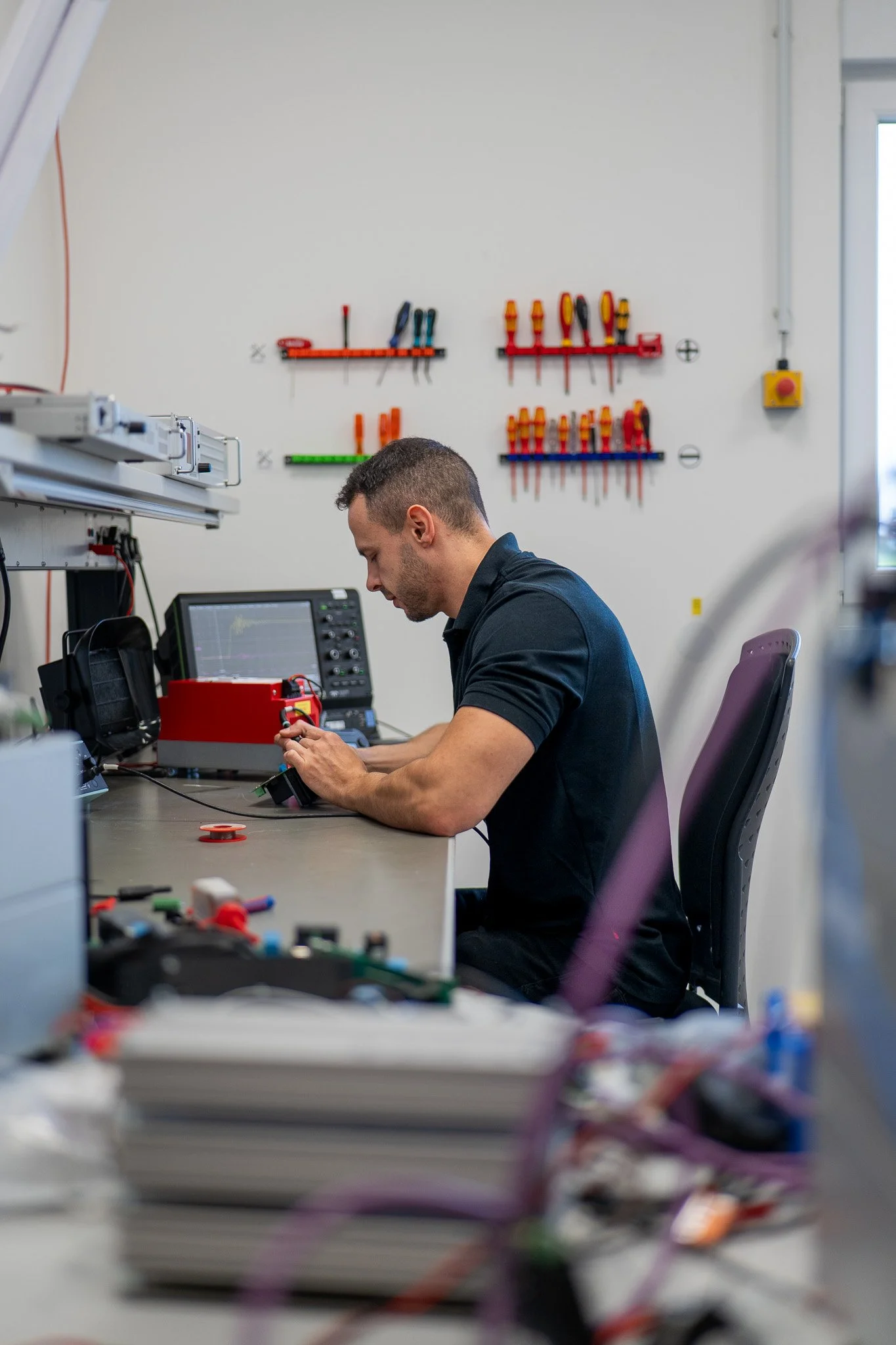 Ein Mann arbeitet in einem technischen Labor an einem Schaltgerät, im Hintergrund Werkzeug an der Wand und auf dem Tisch verteilt.