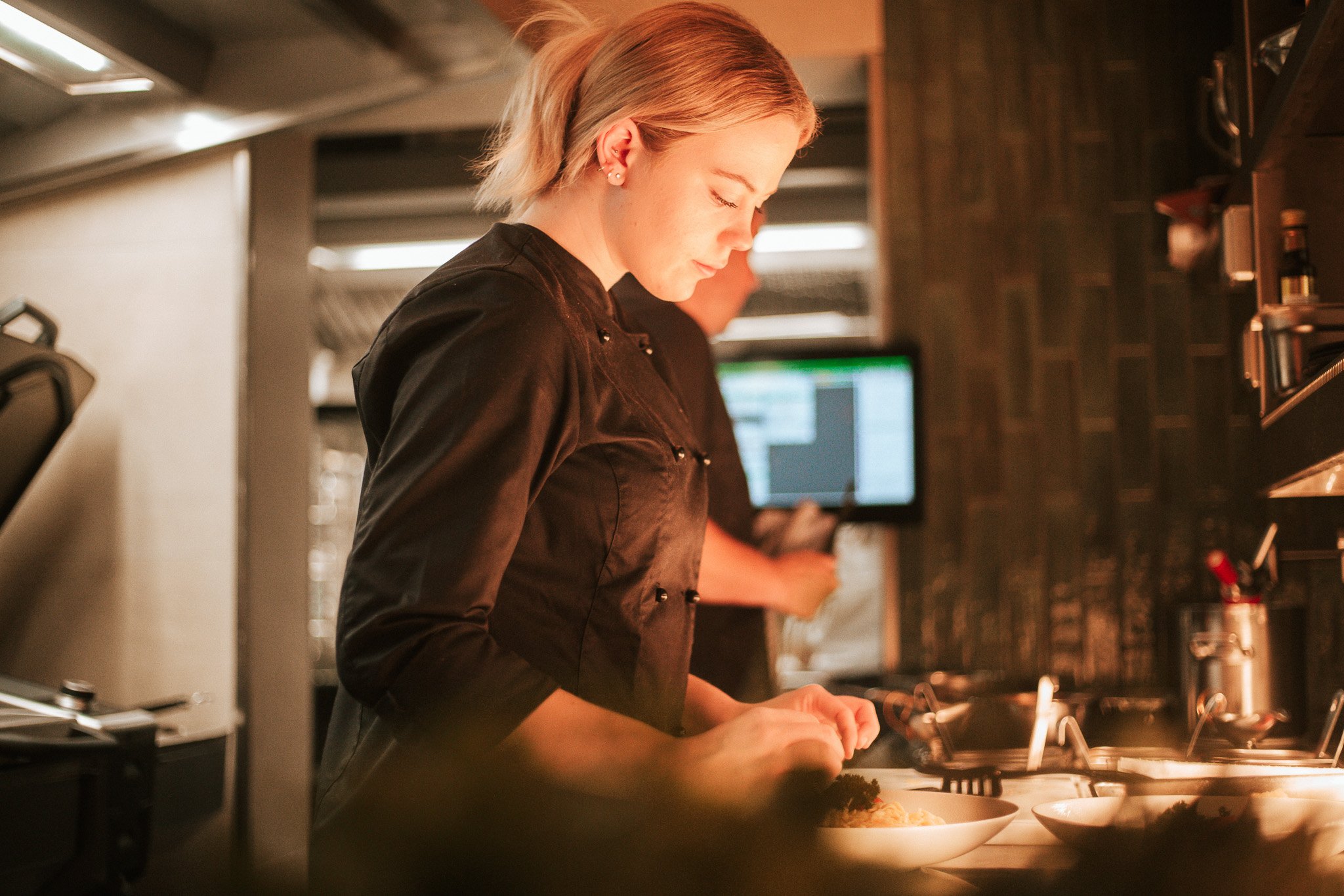 Koch in schwarzer Uniform arbeitet konzentriert in einer Küchenumgebung, vorbereitet Essen auf einem Teller.