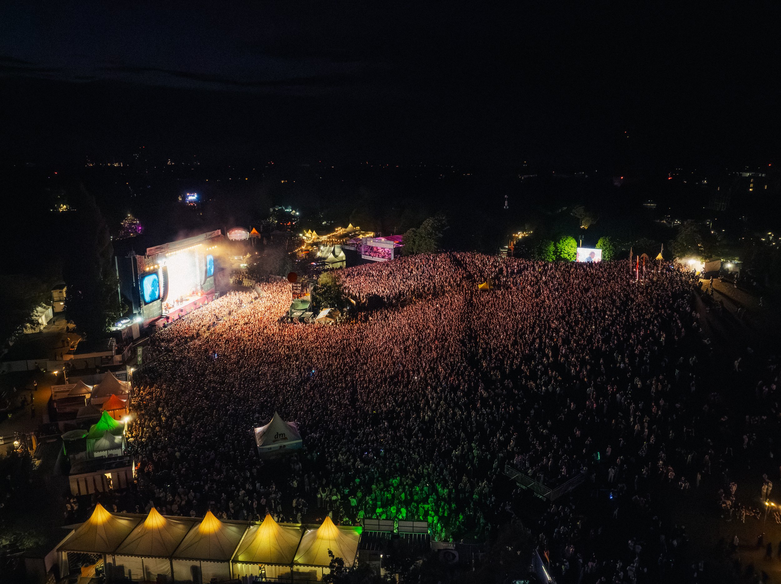 Nachtaufnahme eines großen Konzerts mit einer Menschenmenge vor einer Bühne, umgeben von Zelten und Lichter im Freien.
