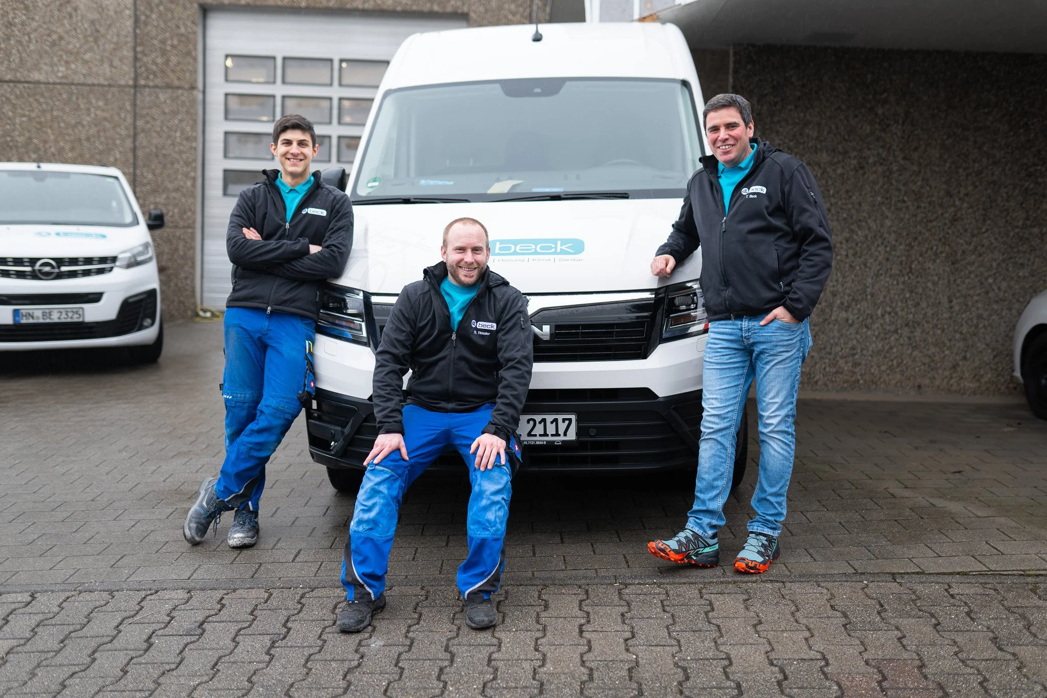 Drei Männer in blauer Arbeitskleidung posieren vor einem weißen Firmenwagen mit "beck"-Logo auf dem Parkplatz eines Gebäudes.