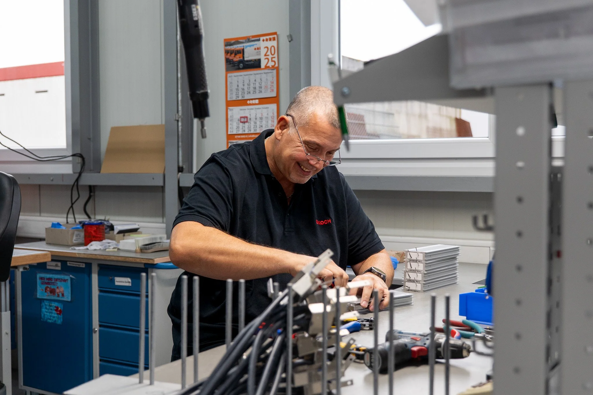 Ein Mann arbeitet in einer Werkstatt an einem Tisch, umgeben von Werkzeugen und kleinen Metallteilen, mit einem Fenster im Hintergrund.