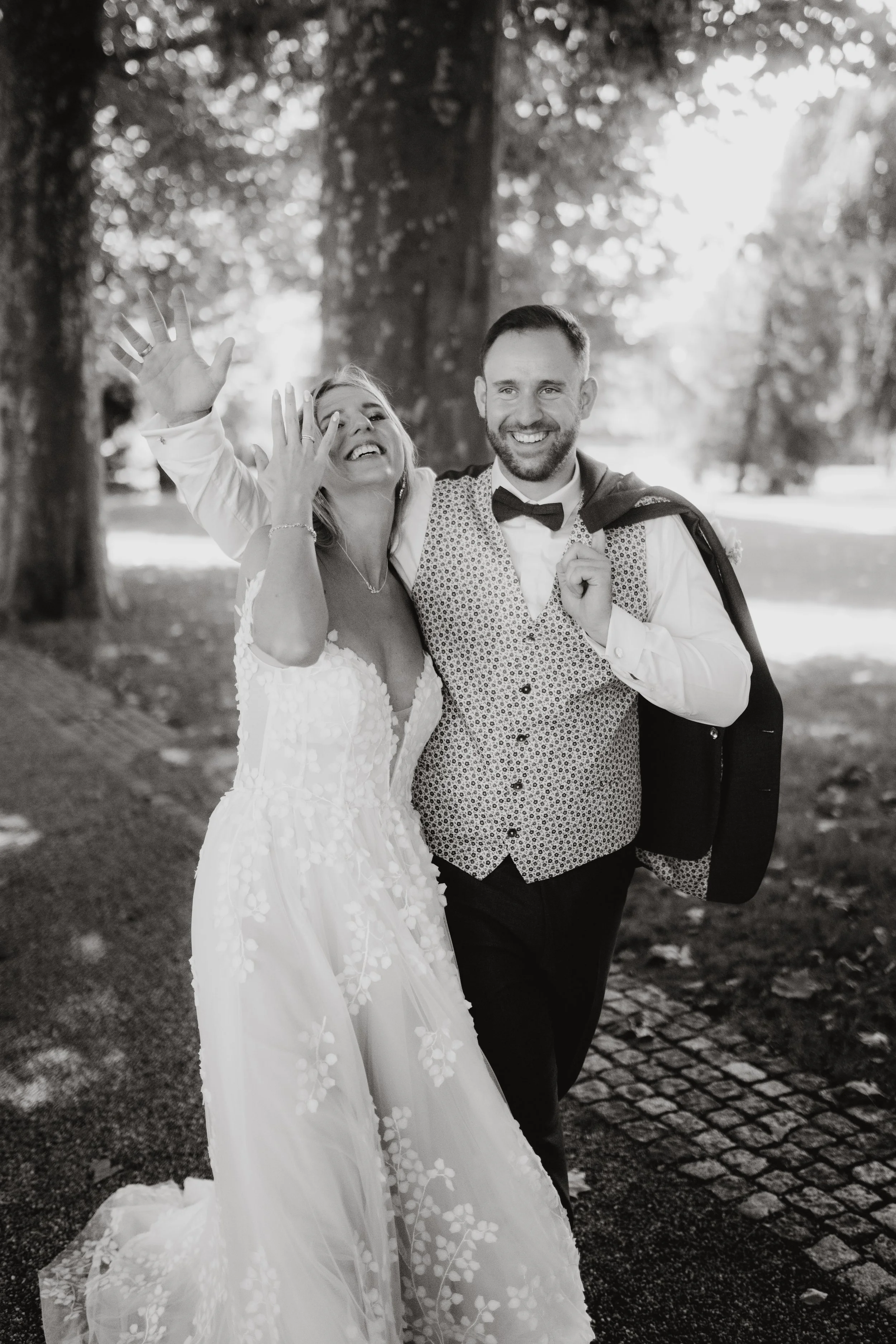 Ein glückliches Paar, vermutlich Braut und Bräutigam, spaziert im Park. Die Frau trägt ein langes Hochzeitskleid, der Mann einen Anzug mit Weste und Krawatte. Beide lachen und haben Händchen gehalten, während sie durch den Park gehen.