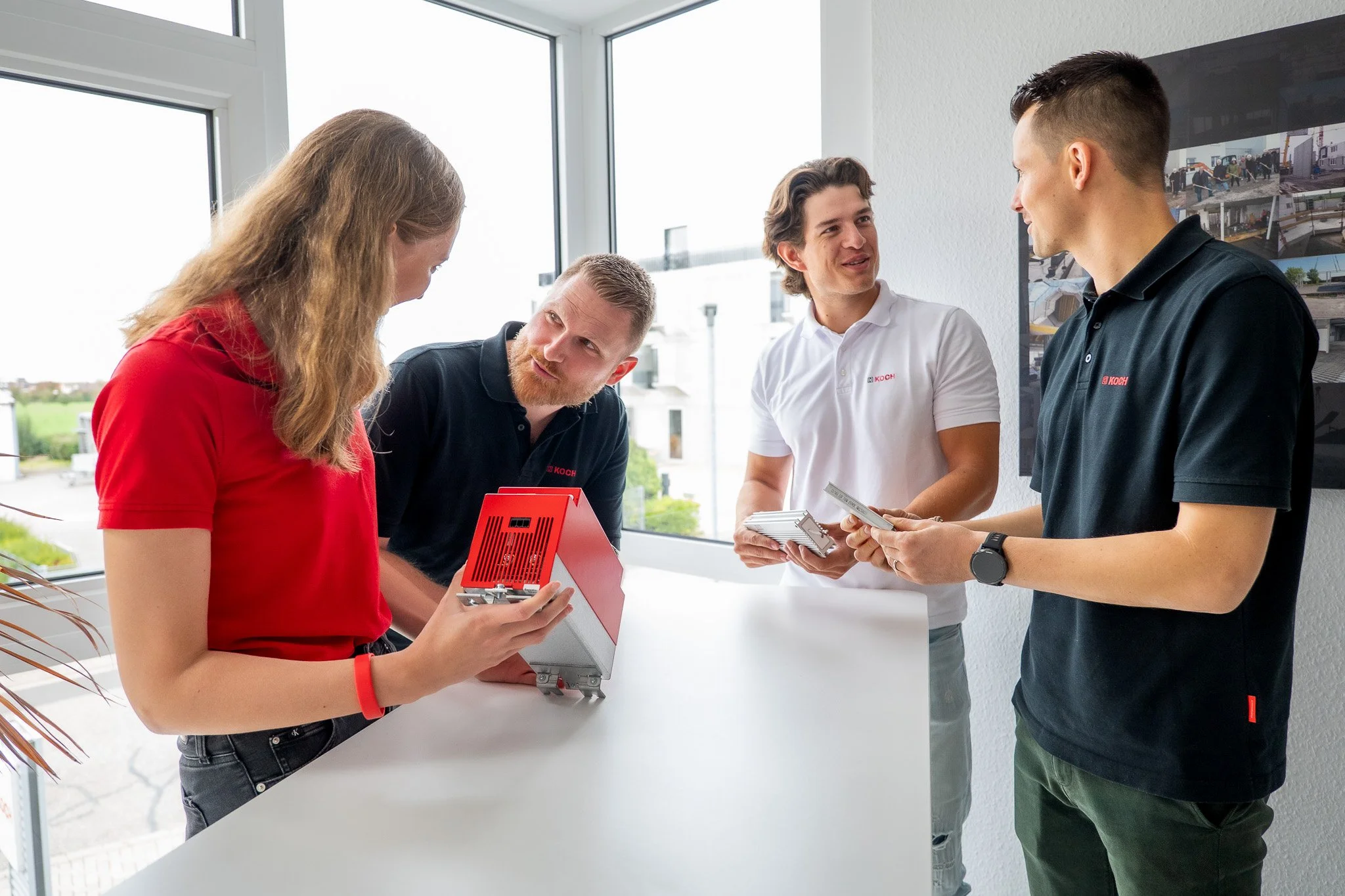 Vier junge Menschen im Gespräch, eine Frau zeigt auf ein rotes Gerät, die anderen drei hören zu und halten Broschüren, im Hintergrund sind großflächige Fenster und Bilder