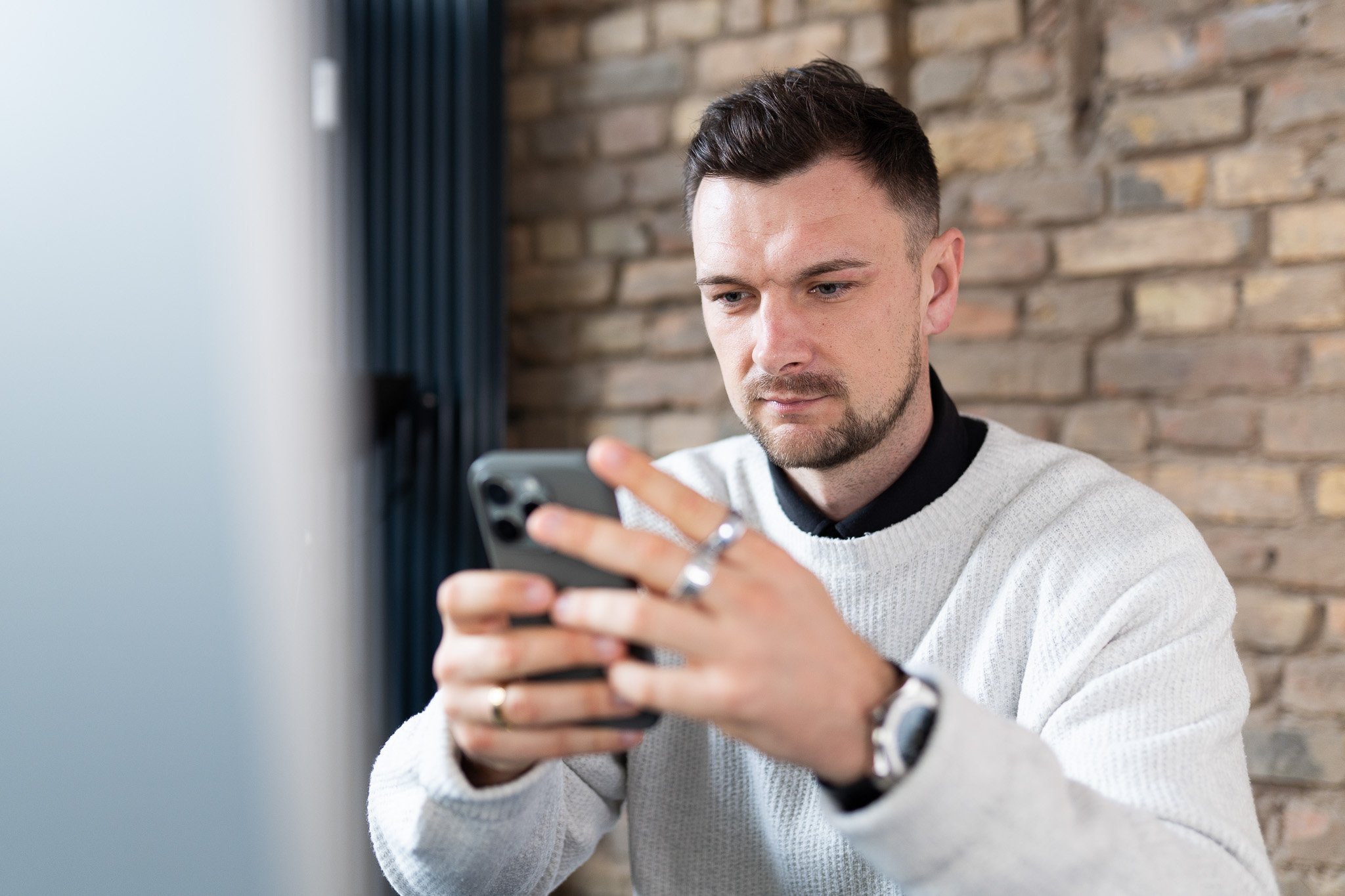 Ein Mann in einem weißen Pullover hält ein Smartphone in der Hand und schaut darauf, vor einer Backsteinwand.