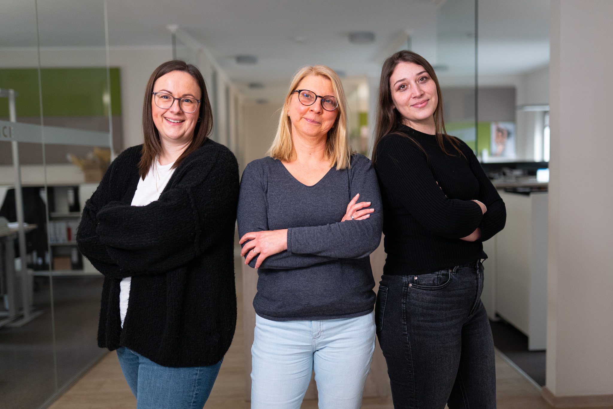 Drei lächelnde Frauen in Freizeitkleidung stehen in einem Büroflur mit den Armen verschränkt.