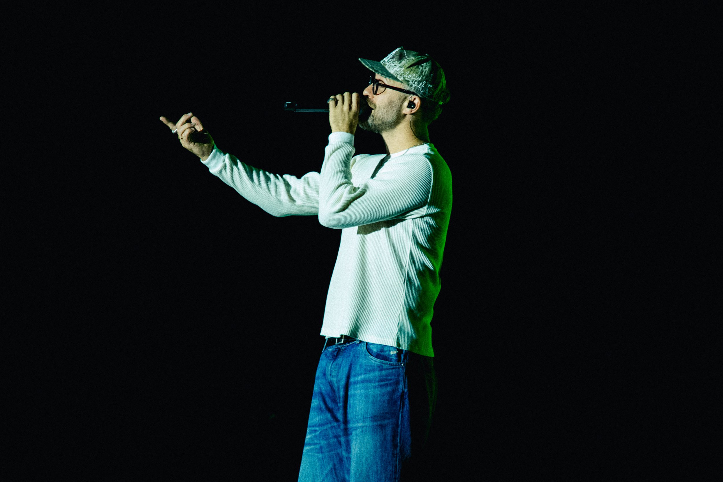 Ein Mann trägt eine Kappe und eine Brille, singt in ein Mikrofon auf einer dunklen Bühne.