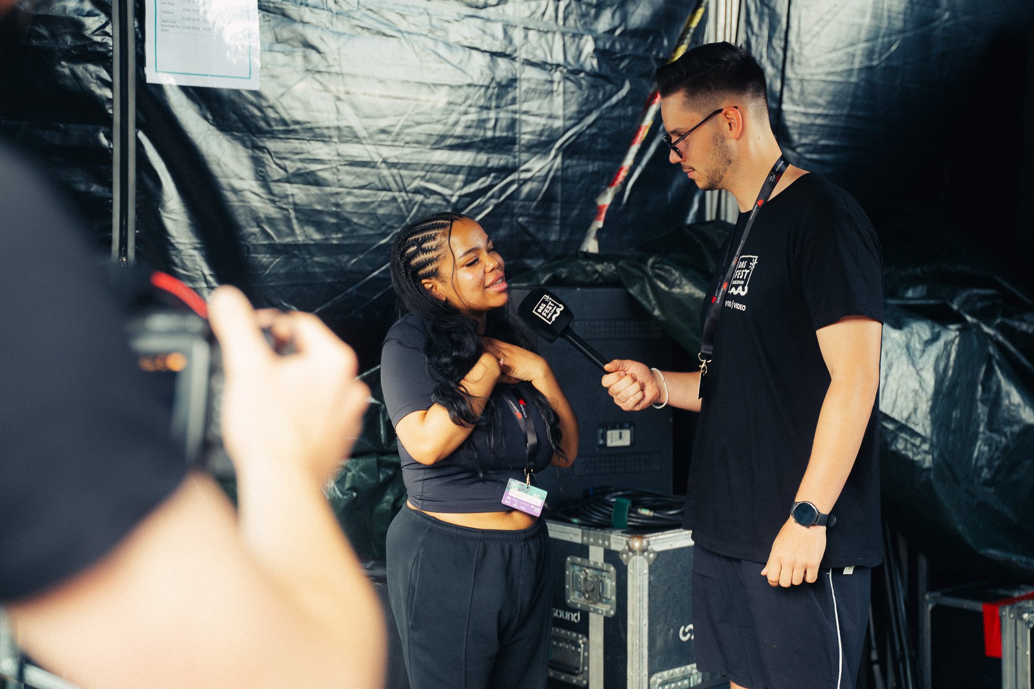Ein Mann interviewt eine lächelnde Frau, während er einen Mikrofon hält, hinter ihnen schwarze Plastikplanen und technische Ausrüstung. Ein weiteres Person is im Vordergrund, vermutlich beim Fotografieren.