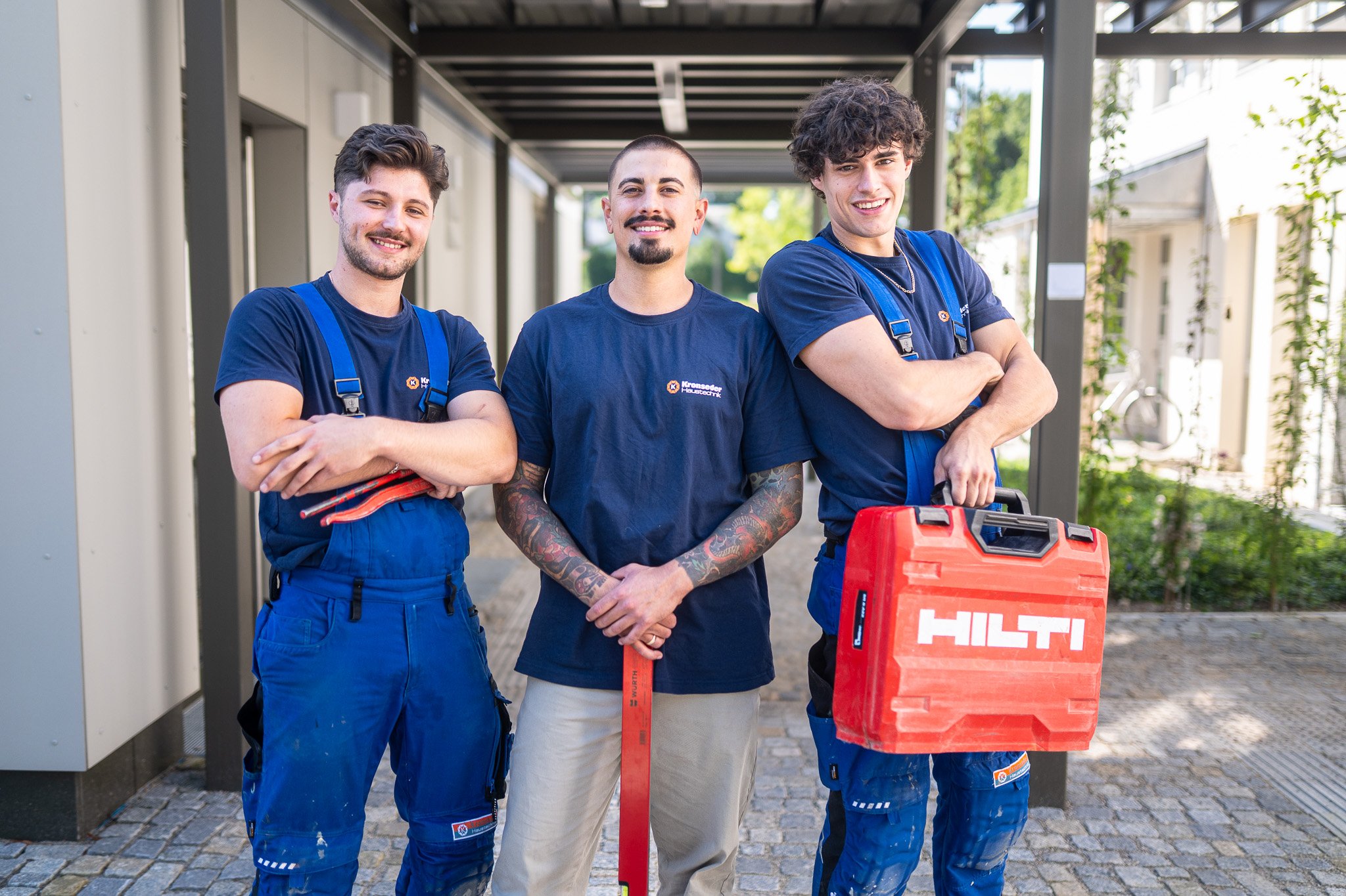 Drei junge Handwerker stehen draußen auf einem Gehweg vor einem Gebäude, lachend und in Arbeitskleidung, mit Werkzeugen und einem roten HILTI-Koffer.