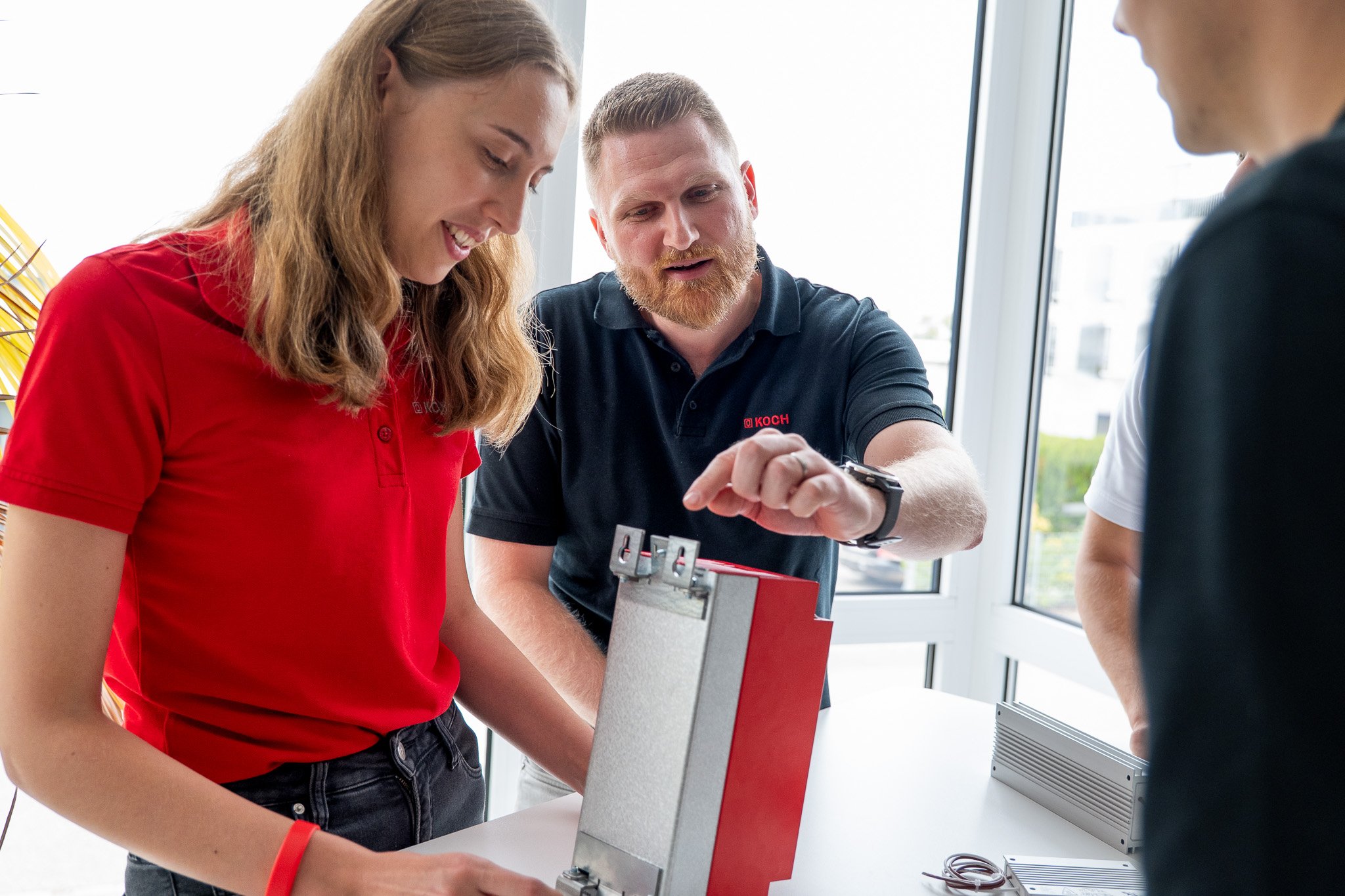 Drei Personen beim technischen Gespräch, eine Person erklärt etwas an einem Gerät, die anderen beiden hören aufmerksam zu.