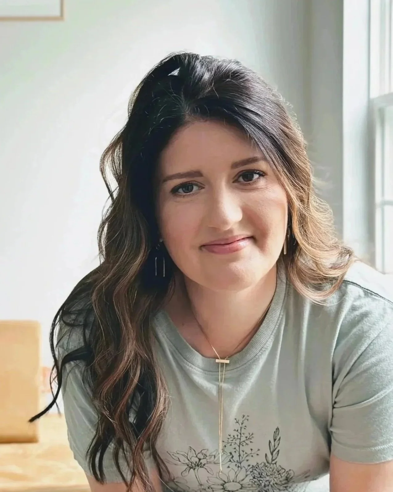 A woman with long wavy brown hair, light skin, and blue eyes smiling, sitting indoors near a window with natural light, wearing a light gray T-shirt with floral print and a delicate gold necklace.