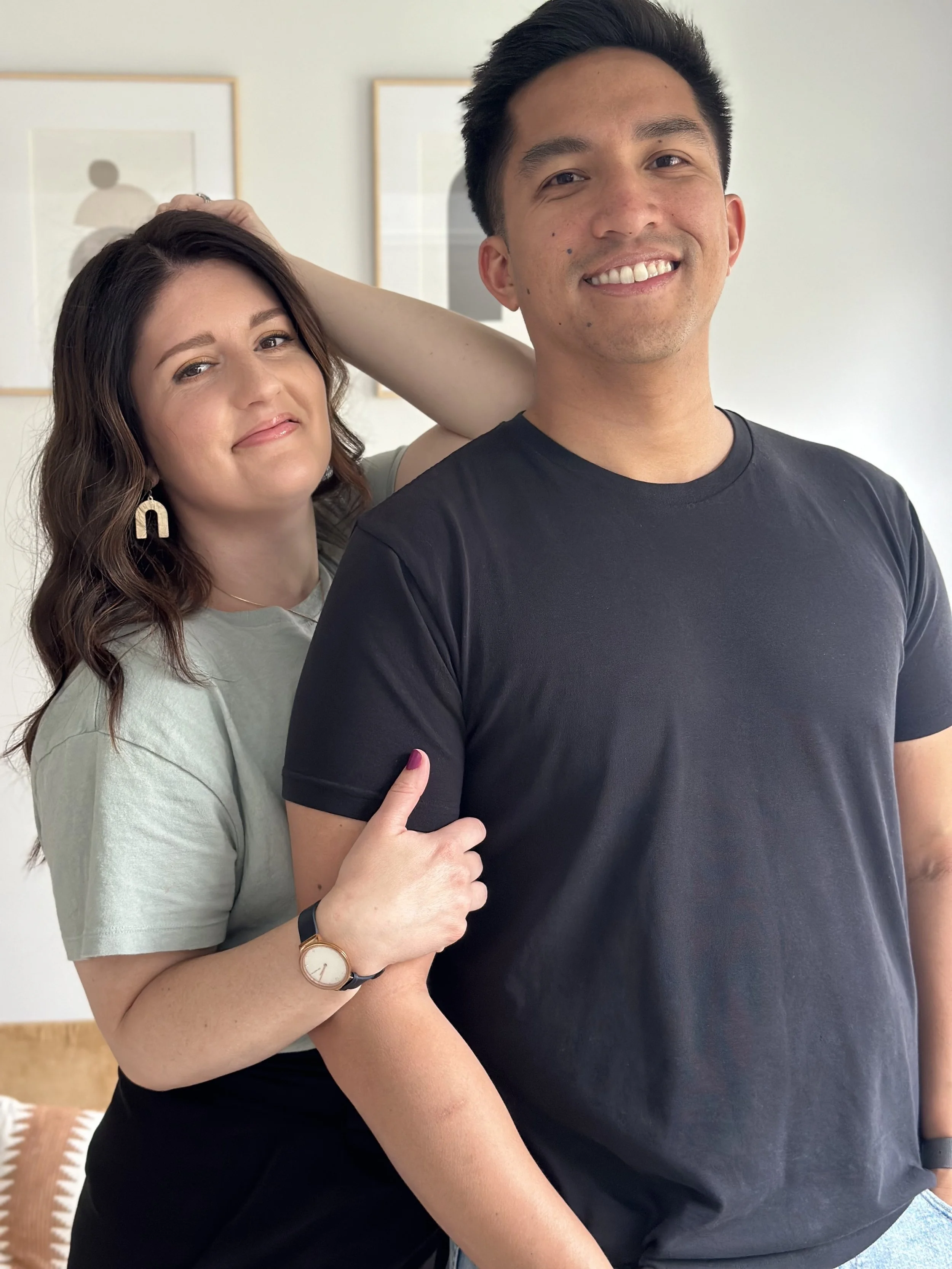 A woman with wavy brown hair and earrings making a peace sign with her hand, smiling, standing behind a man with short black hair, smiling, wearing a black t-shirt, in a room with framed artwork on the wall.