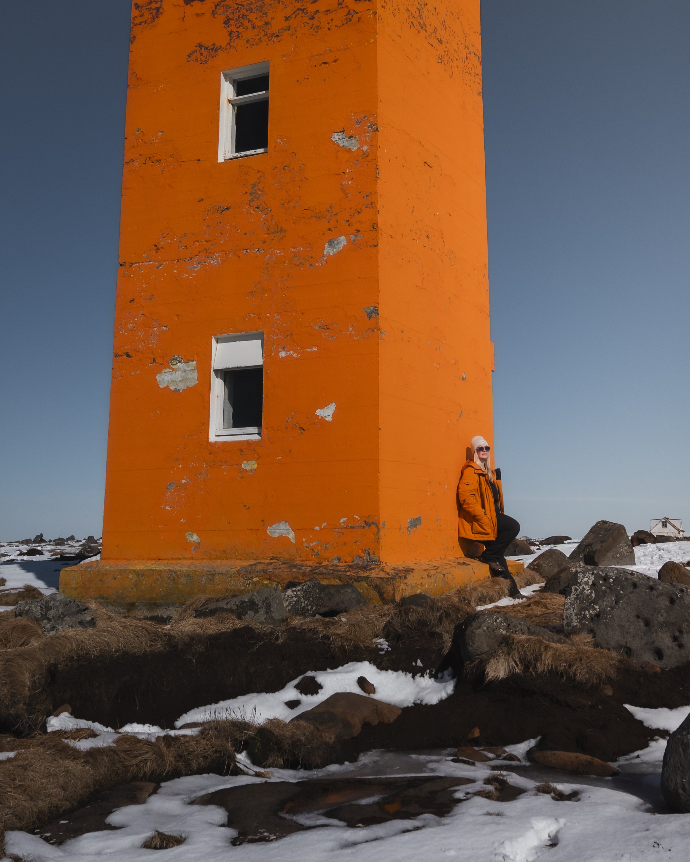 A woman with sunglasses wearing an orange jacket leaning against an orange lighthouse in a snowy, rocky landscape.