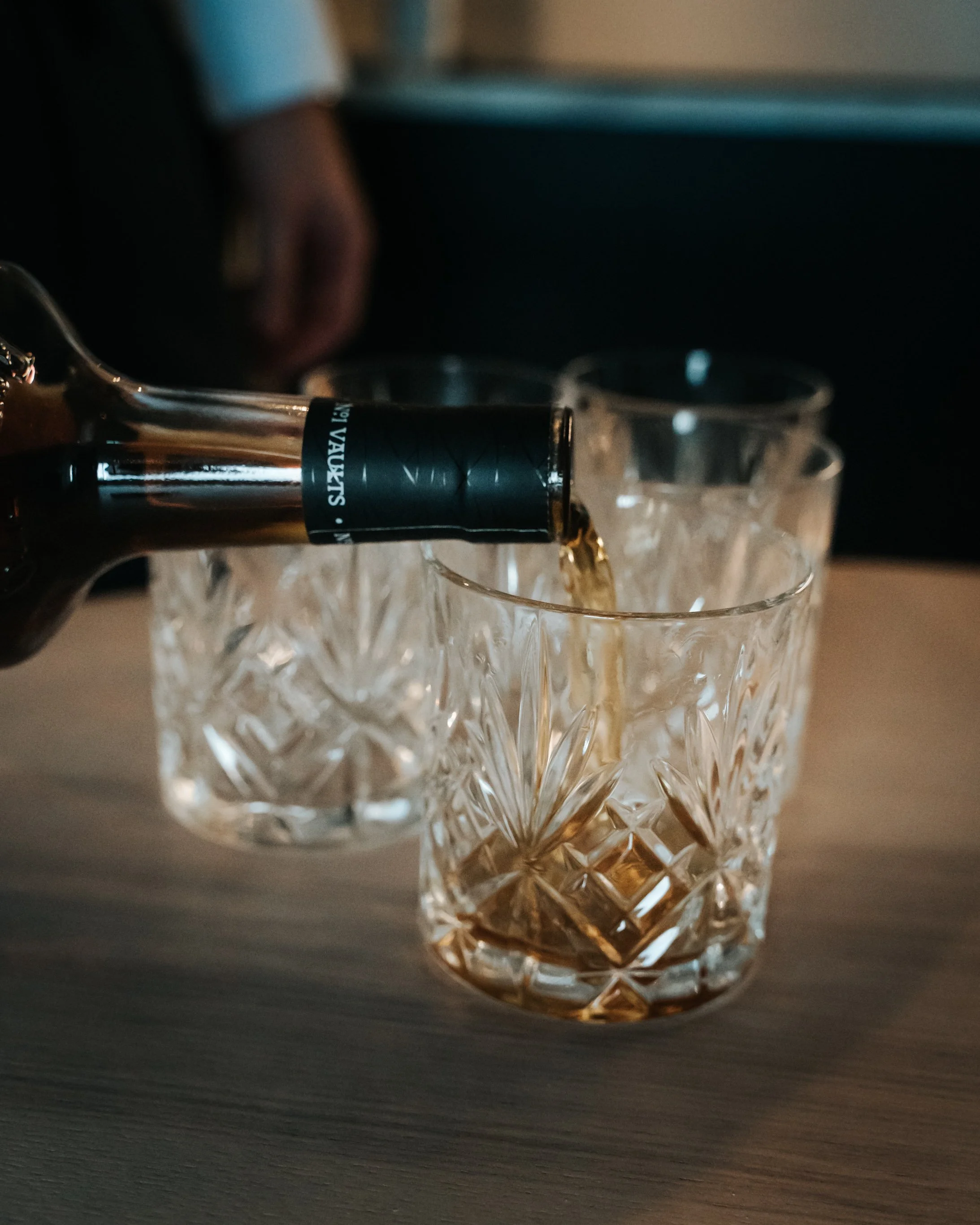 A hand pouring whiskey from a bottle into a cut glass tumbler on a wooden surface.