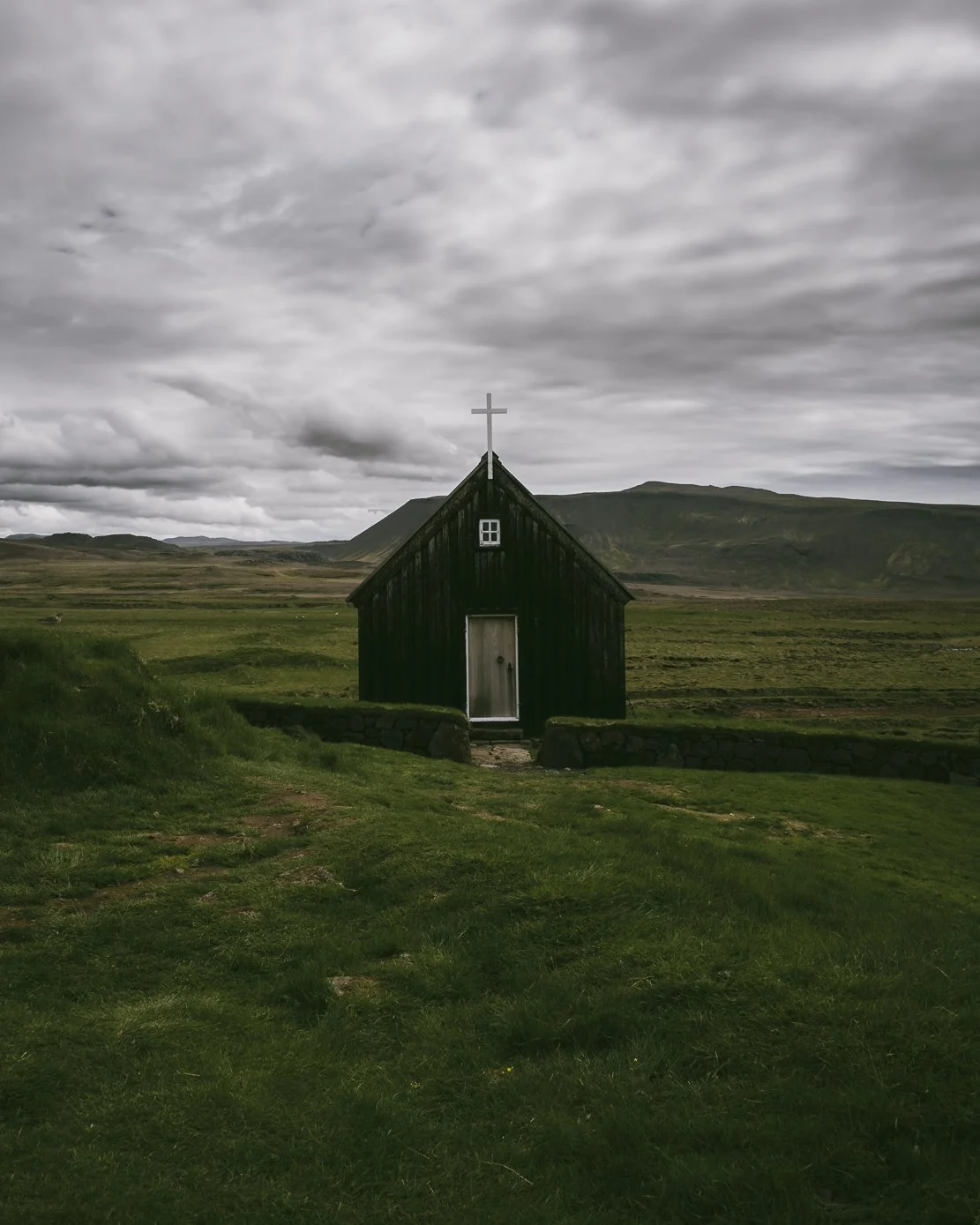 A small black wooden church with a white cross on top, situated in a green field with rolling hills and a cloudy sky in the background.