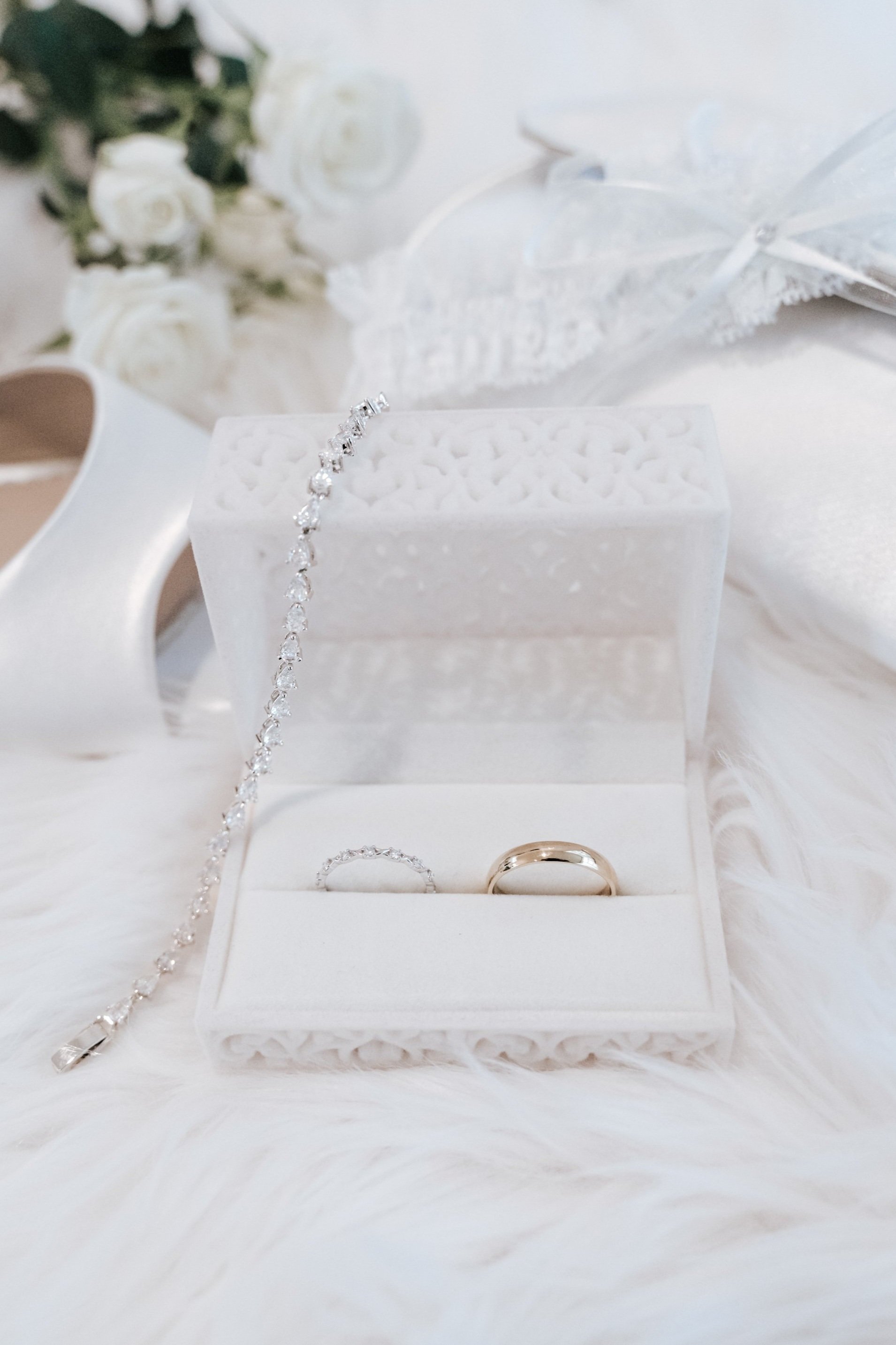 A white jewelry box containing a diamond ring and a wedding band, with a diamond bracelet and white roses in the background, on a white fluffy surface.